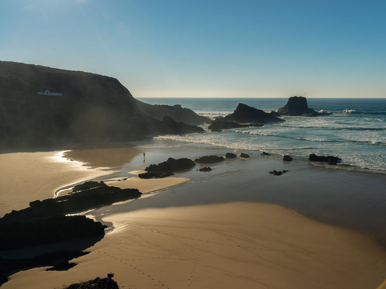 Vista aérea de una playa desierta con acantilados rocosos, mar agitado y una persona caminando bajo un cielo azul