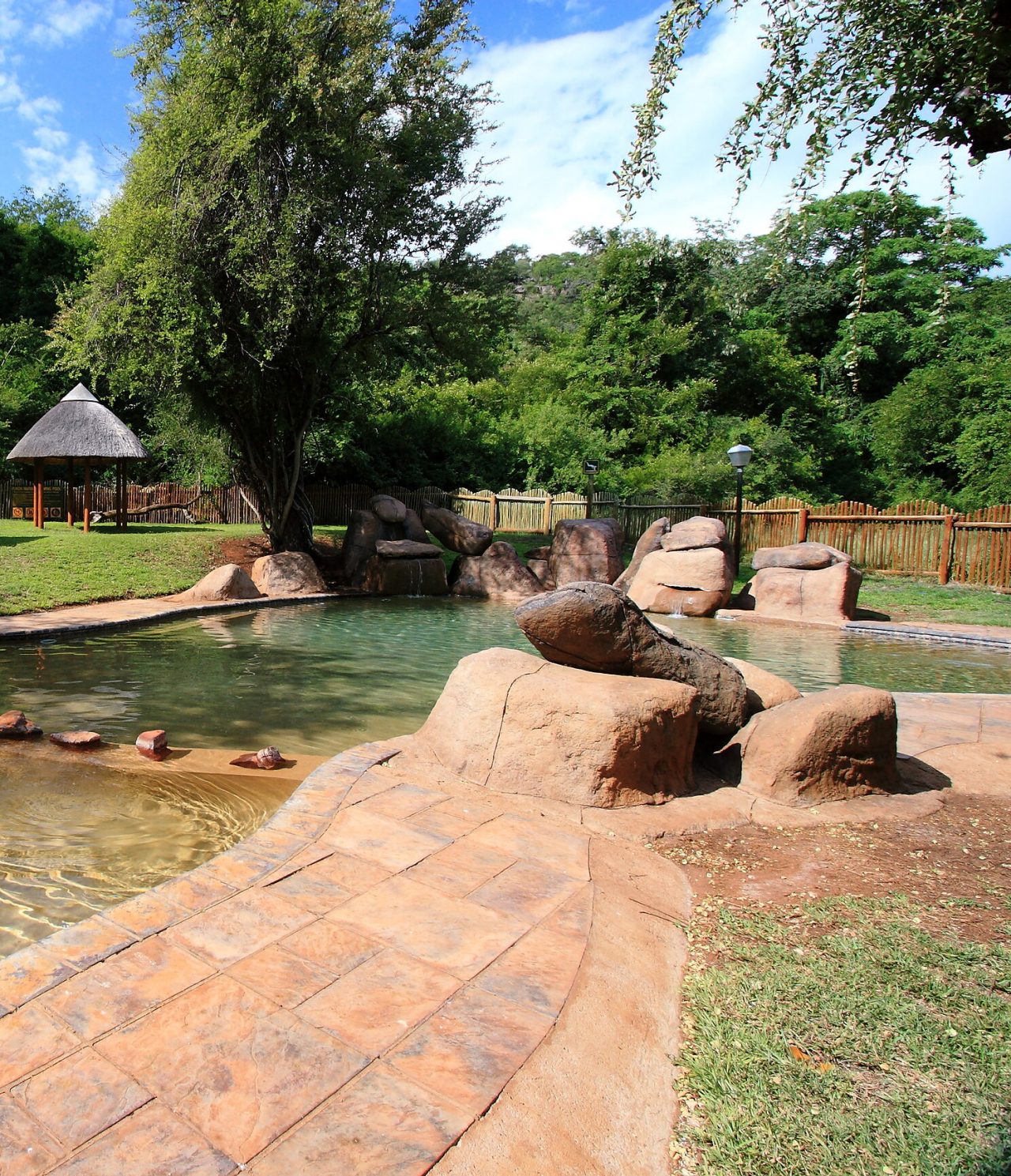 Piscina artificial Kruger Park, com pedras e água verde, rodeada por vegetação verdejante e com uma cabana ao fundo