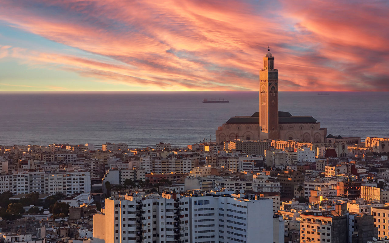 Vista aérea sobre Casablanca, com a luz do pôr-do-sol a iluminar os vários prédios, o céu no horizonte sobre o mar e a torre