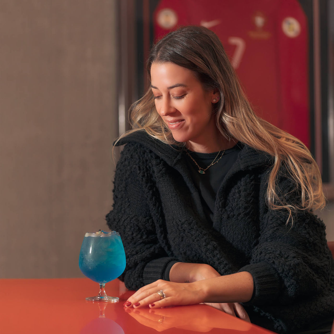 Rapariga a beber um cocktail, sentada numa mesa, com a camisola do Cristiano Ronaldo pendurada na parede