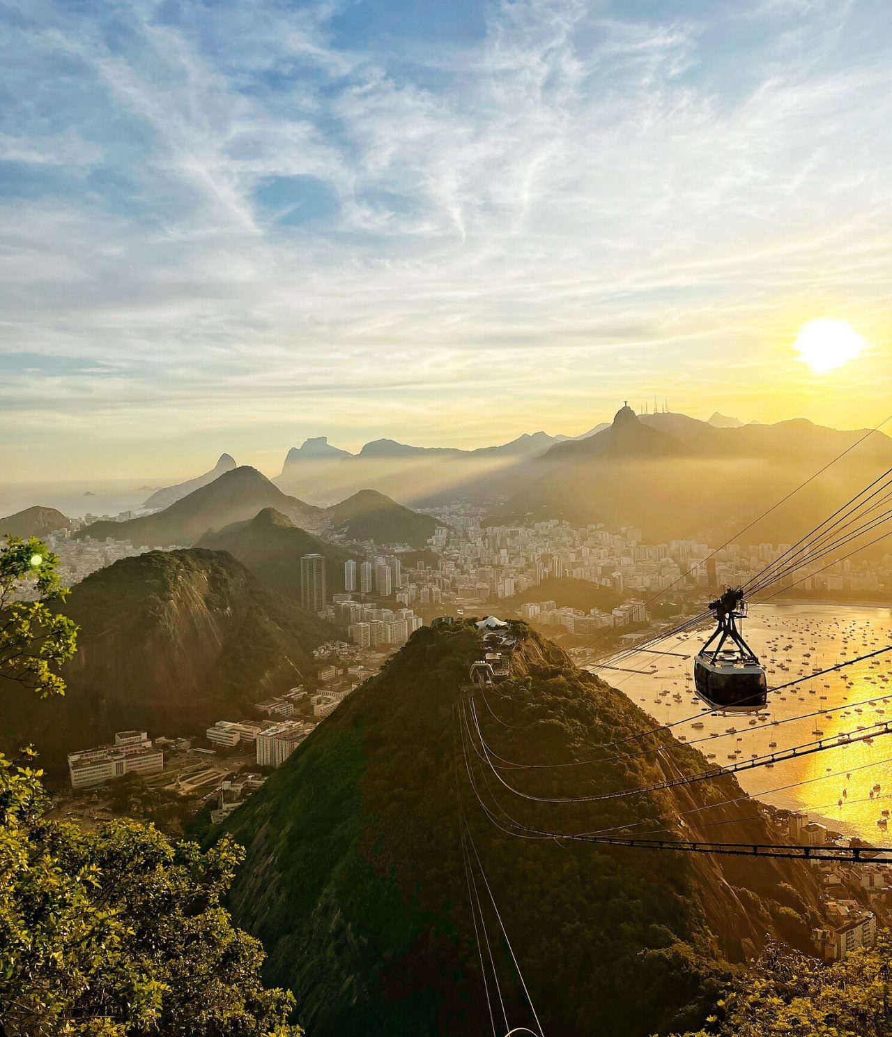 Vista aérea do Rio de Janeiro, com um teleférico a ir em direção a uma colina, com a cidade e a praia no fundo