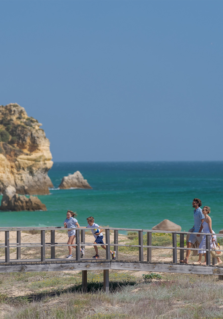 Passadiços de madeira das praias do Alvor, com o mar no fundo e duas crianças a correr com os pais atrás