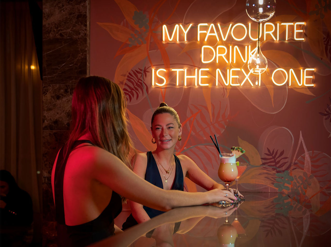 O bar The Bridge, do Hotel 4 Estrelas em Londres, tem duas mulheres no balcão com cocktails e uma frase néon na parede