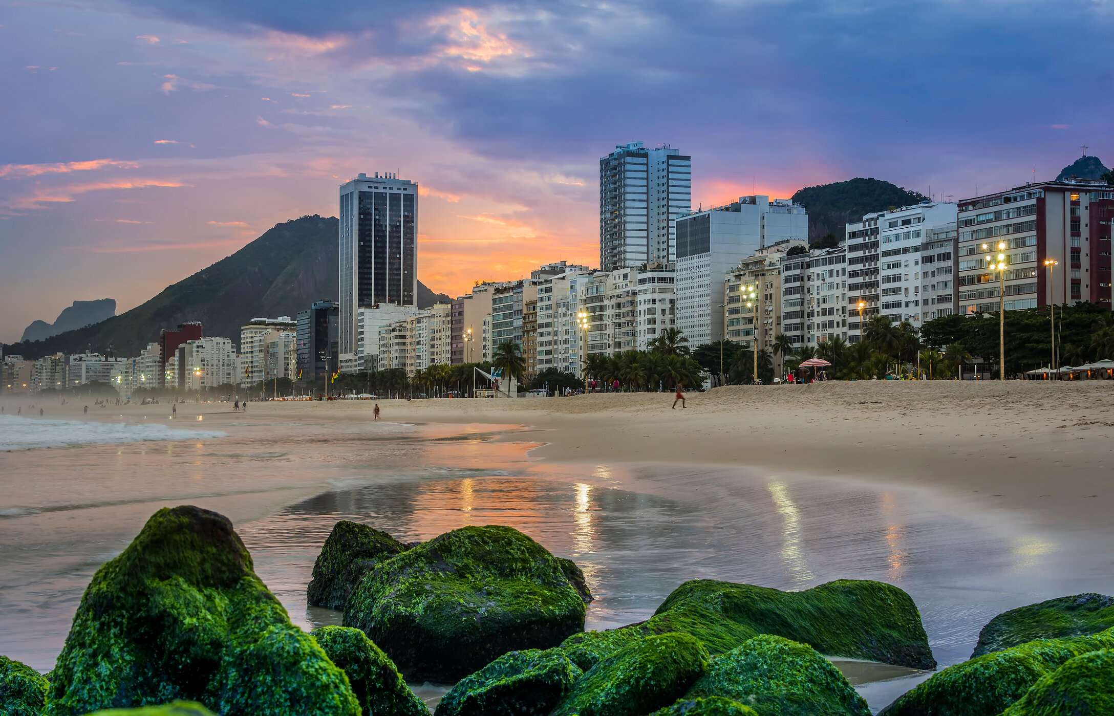 Copacabana-Strand, mit goldenem Sand, Meereswellen und einer belebten Promenade, umgeben von Gebäuden und Bergen im Hintergrund