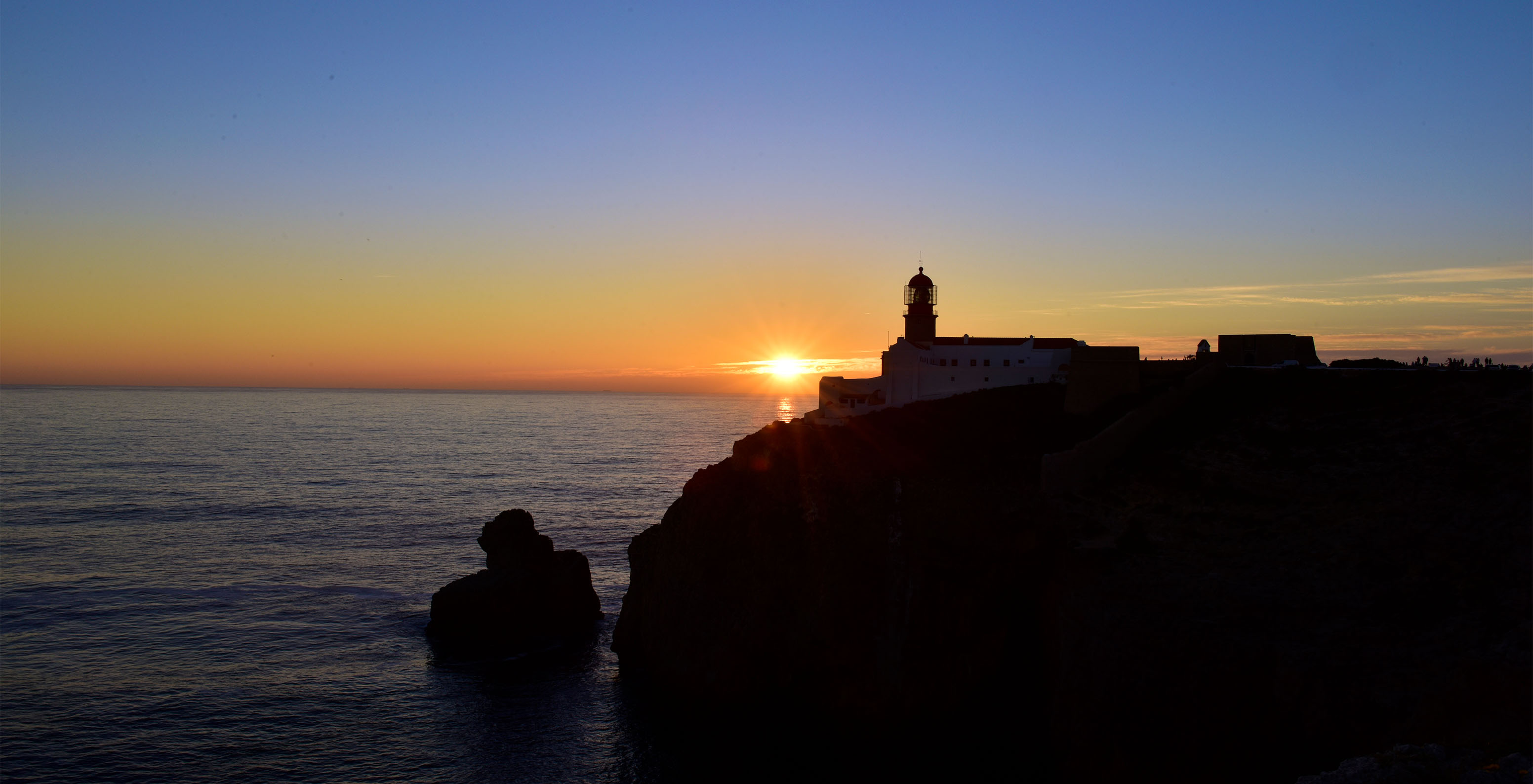 Sonnenuntergang auf einer Klippe mit orangefarbenem Horizont und einem Leuchtturm oben auf der Klippe