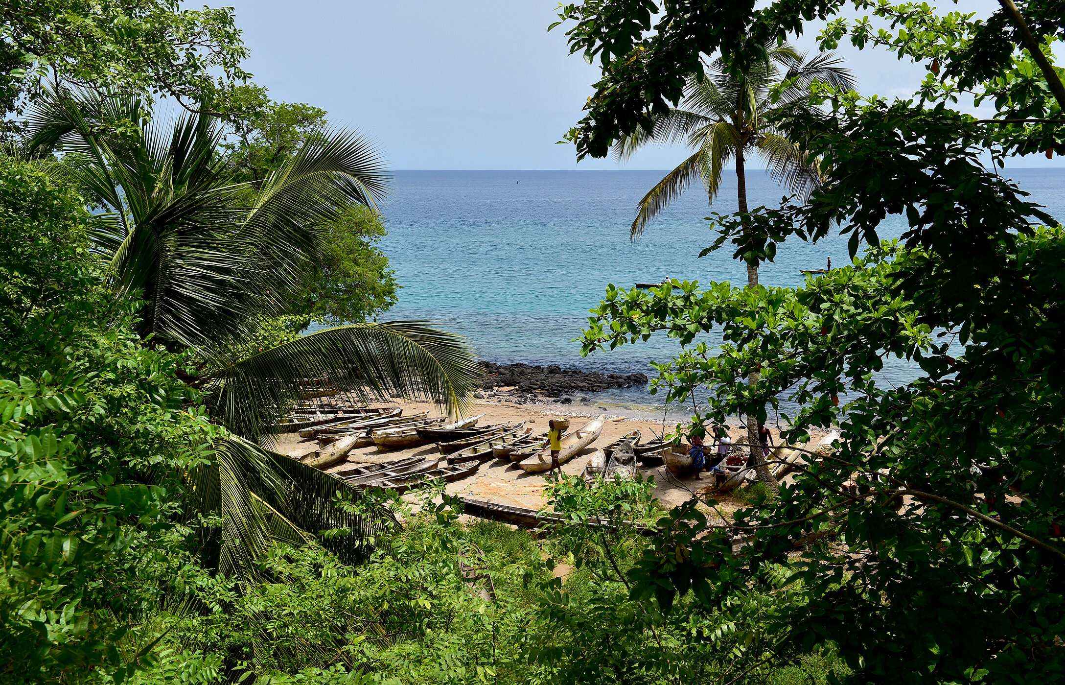 Blick auf einen Strand in São Tomé und Príncipe, umgeben von Vegetation, mit Kanus auf dem Sand und dem Meer im Hintergrund.