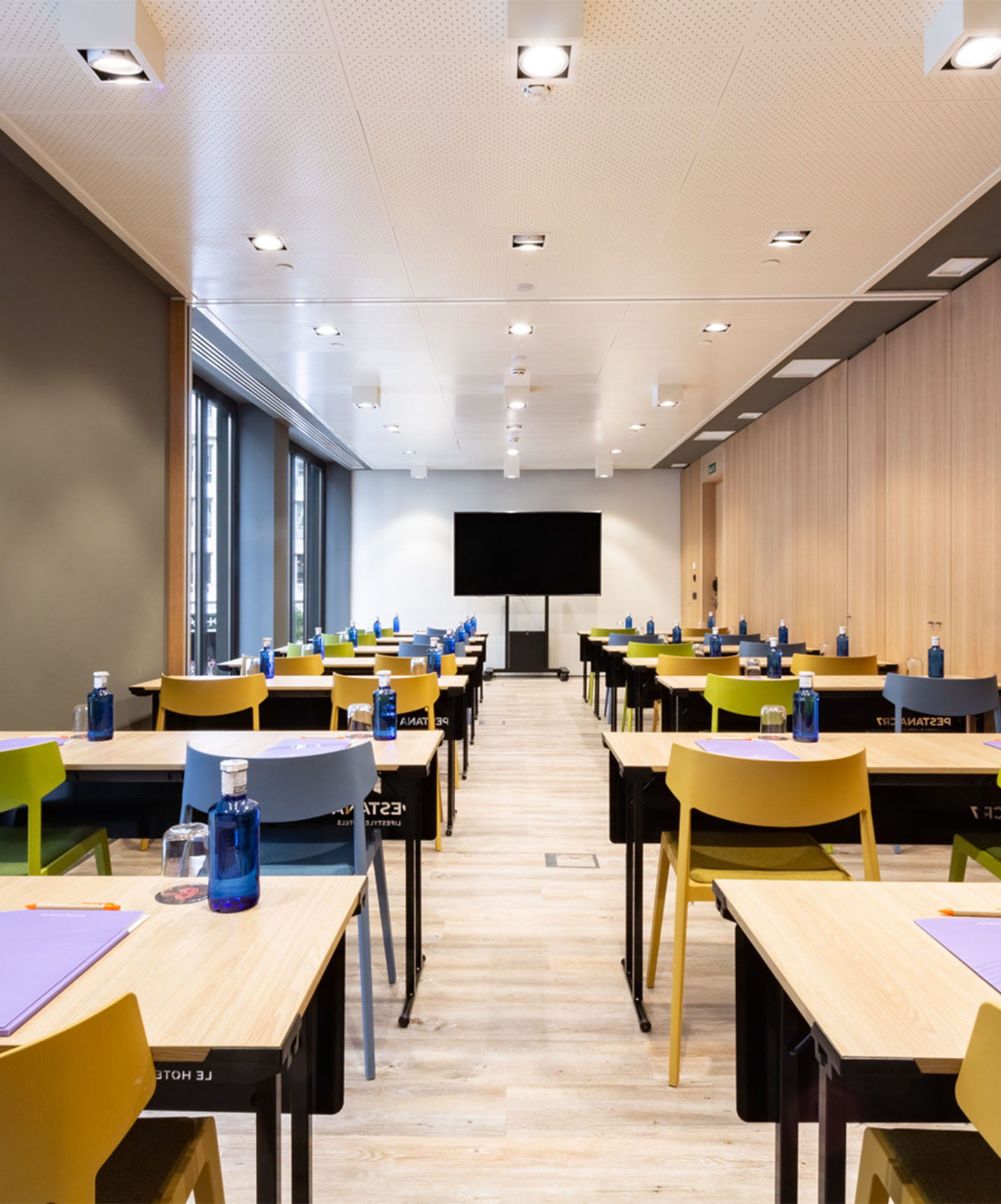 Veranstaltungs- und Besprechungsraum mit Holztischen und Stühlen in Gelb, Grün und Blau, mit Fernseher und Holzverkleidungen