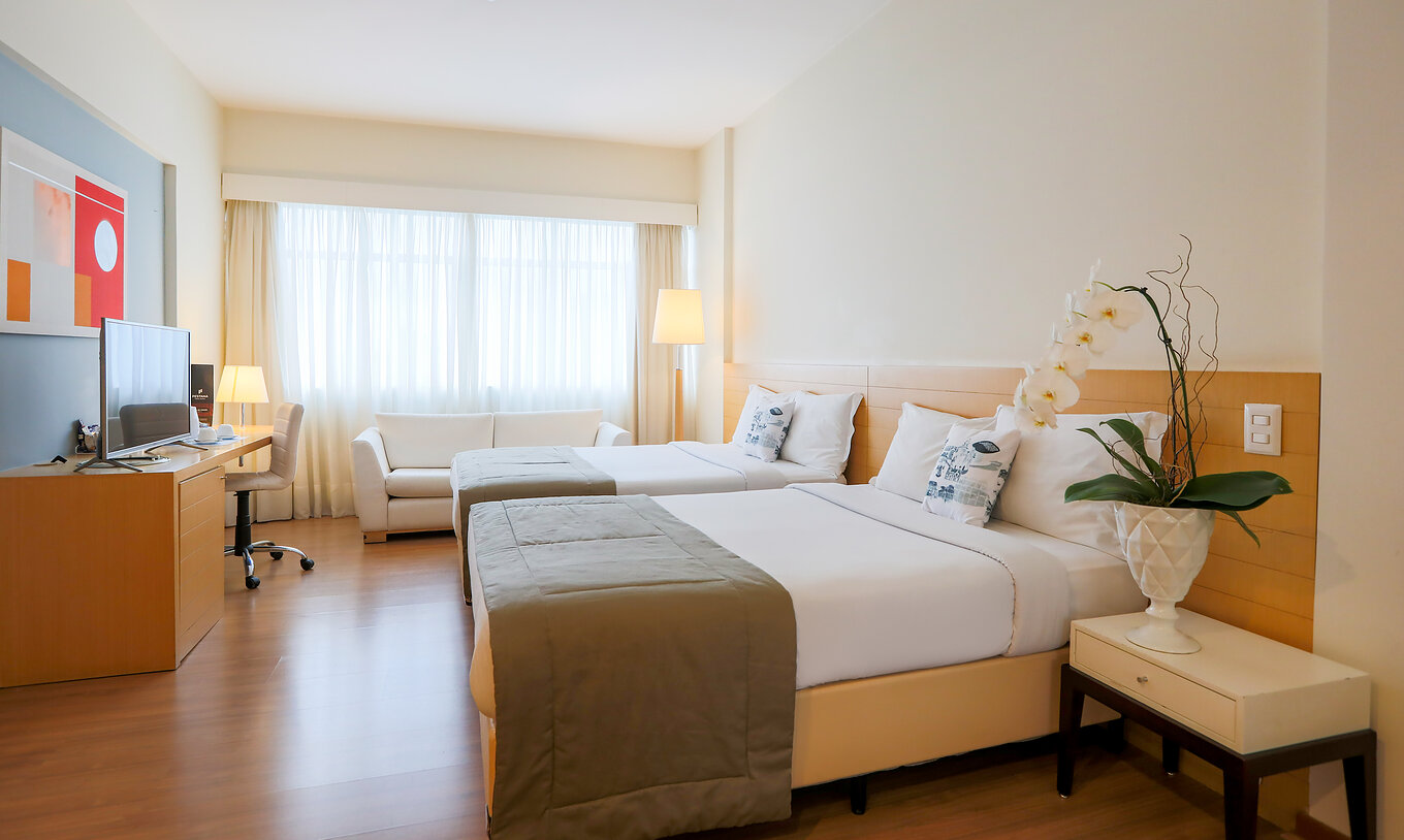 The Family Room at Pestana Curitiba has two double beds, a desk with a television, and a double sofa by the window