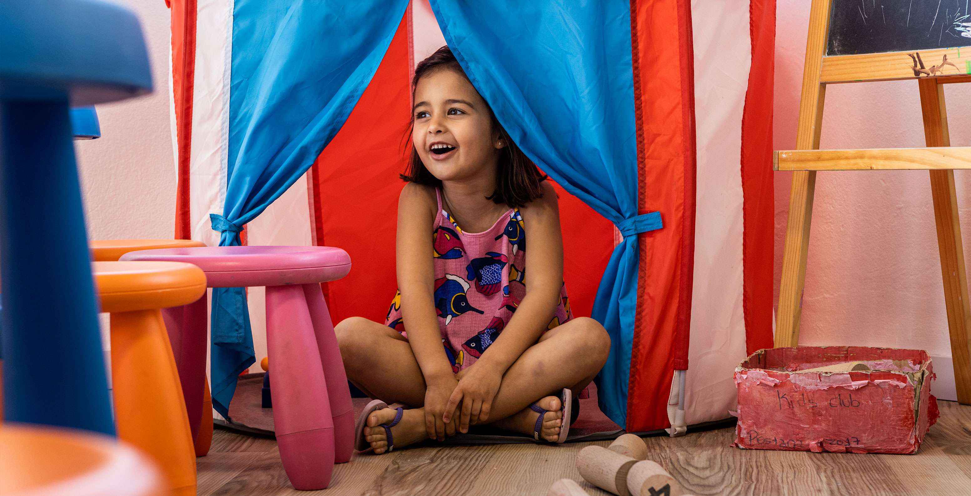 A smiling child plays at the Kids Club, hiding in a red and blue tent