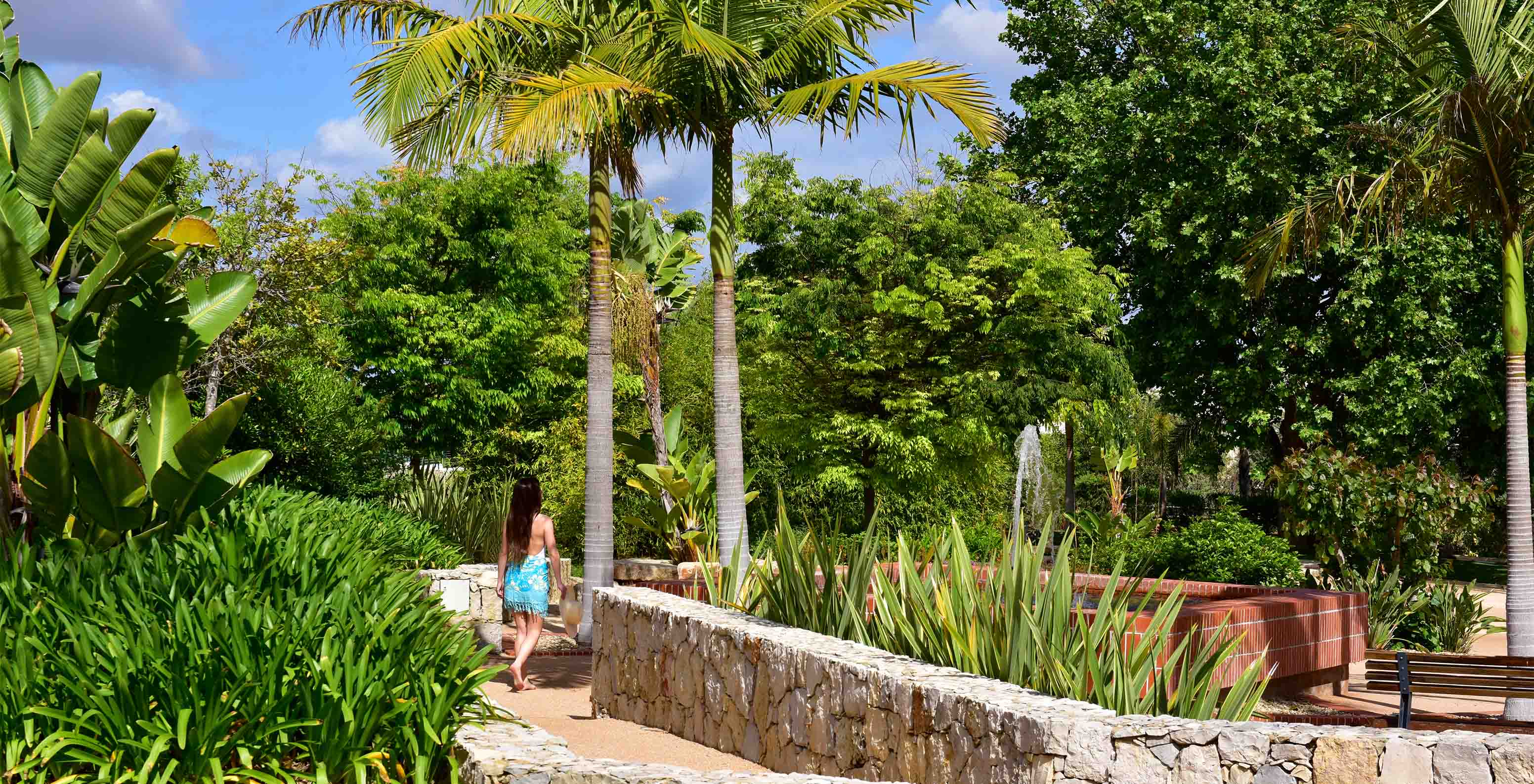 Garden with grass, palm trees, various trees, woman on a boardwalk with stone walls