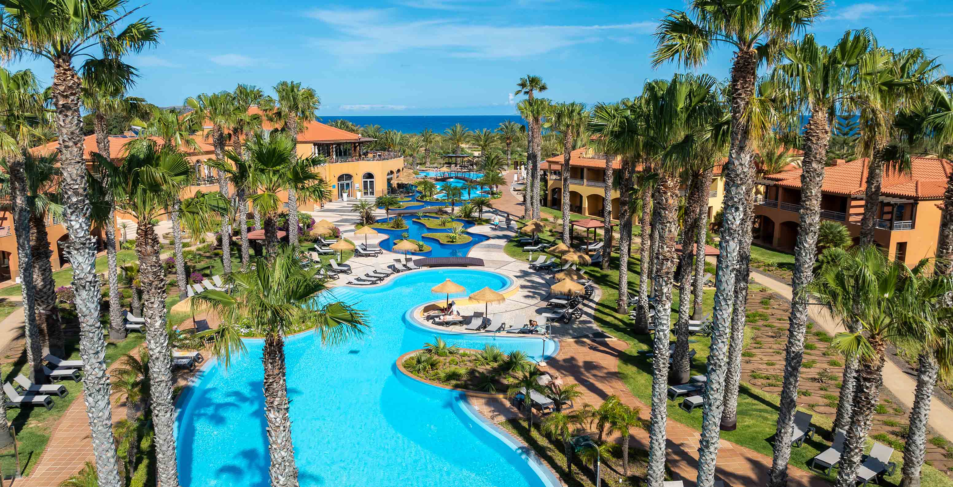 View of the pool at Pestana Porto Santo surrounded by palm trees and colorful buildings under a blue sky