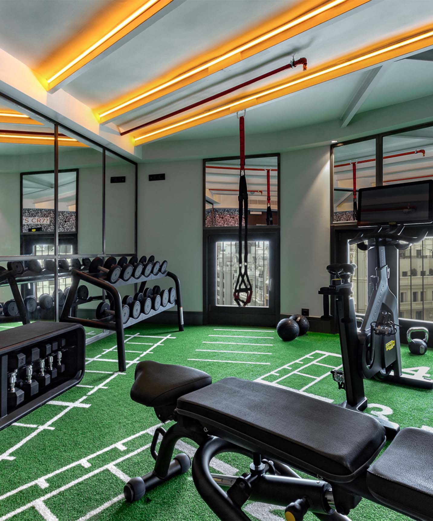 Gym with dumbbells, bike, weight bench, and TRX with mirrors and large windows overlooking front buildings