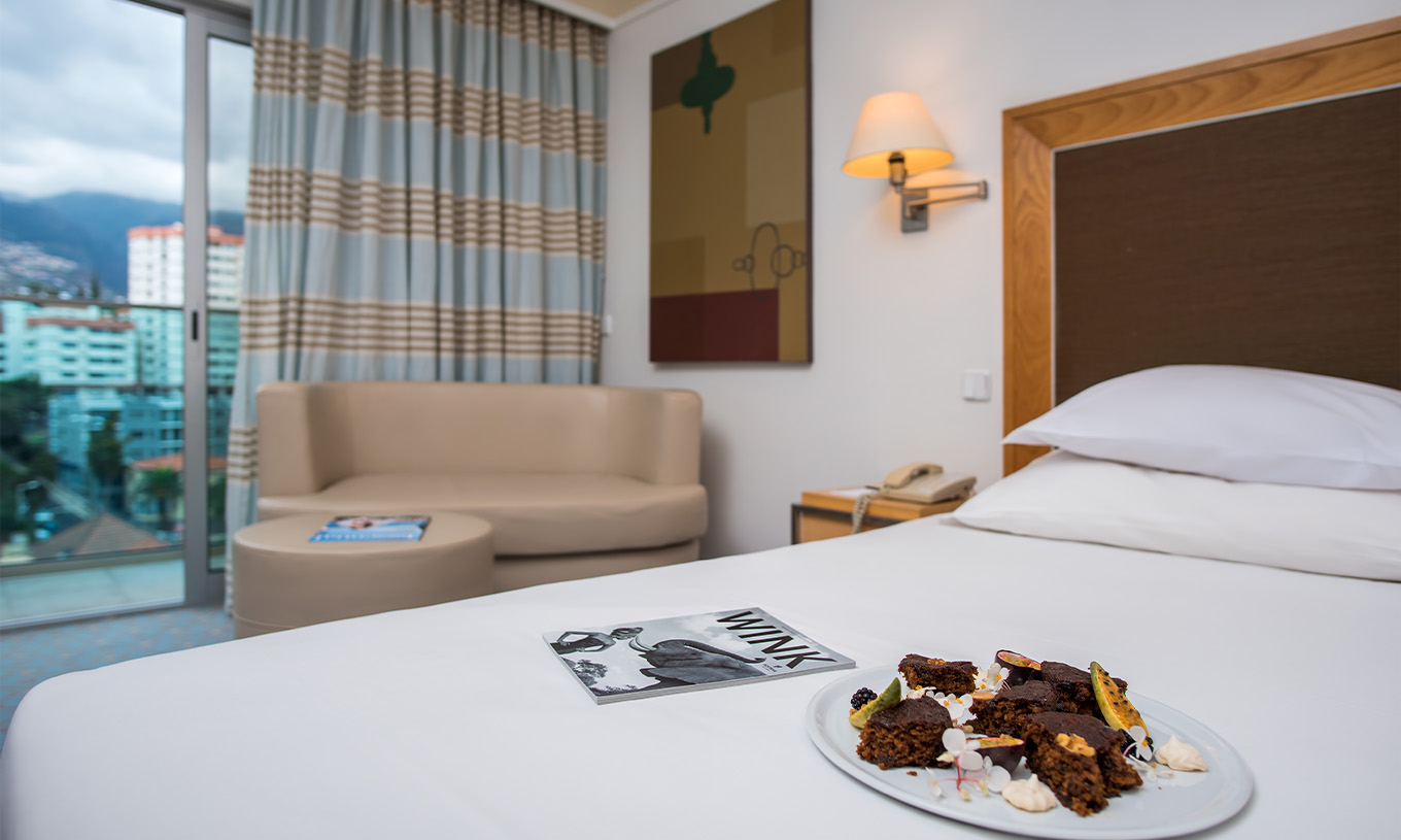 The Twin Classic Room with Bay and Sea View at Pestana Carlton Madeira has a bed with a plate of chocolate and fruit on it