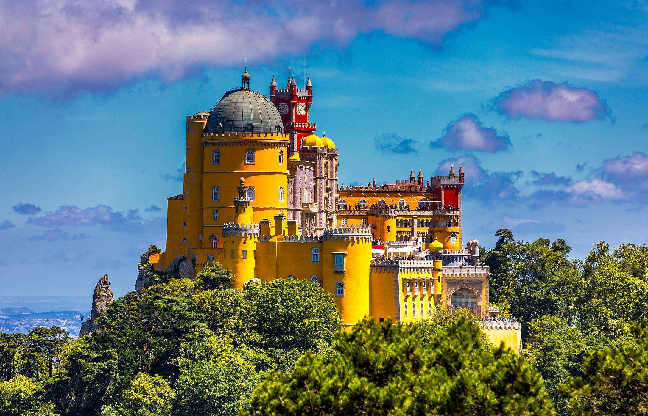 Aerial view of Pena Palace in Sintra, with its vibrant colors, surrounded by vegetation under a blue sky.