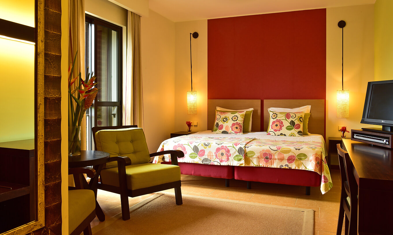 The Junior Suite of Pestana São Tomé is a spacious room with two single beds together, an armchair, and a desk with TV