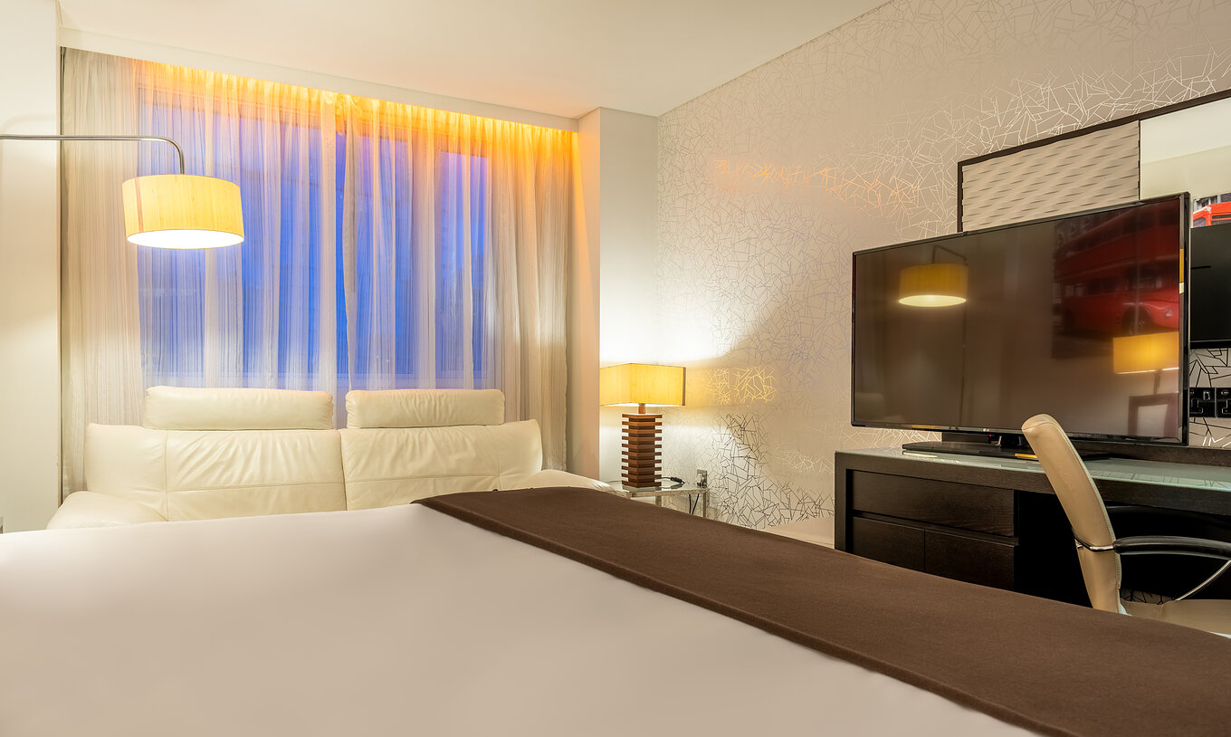 The Deluxe King at Pestana Chelsea Bridge has bed, desk with TV and chair, sofa and windows facing the city