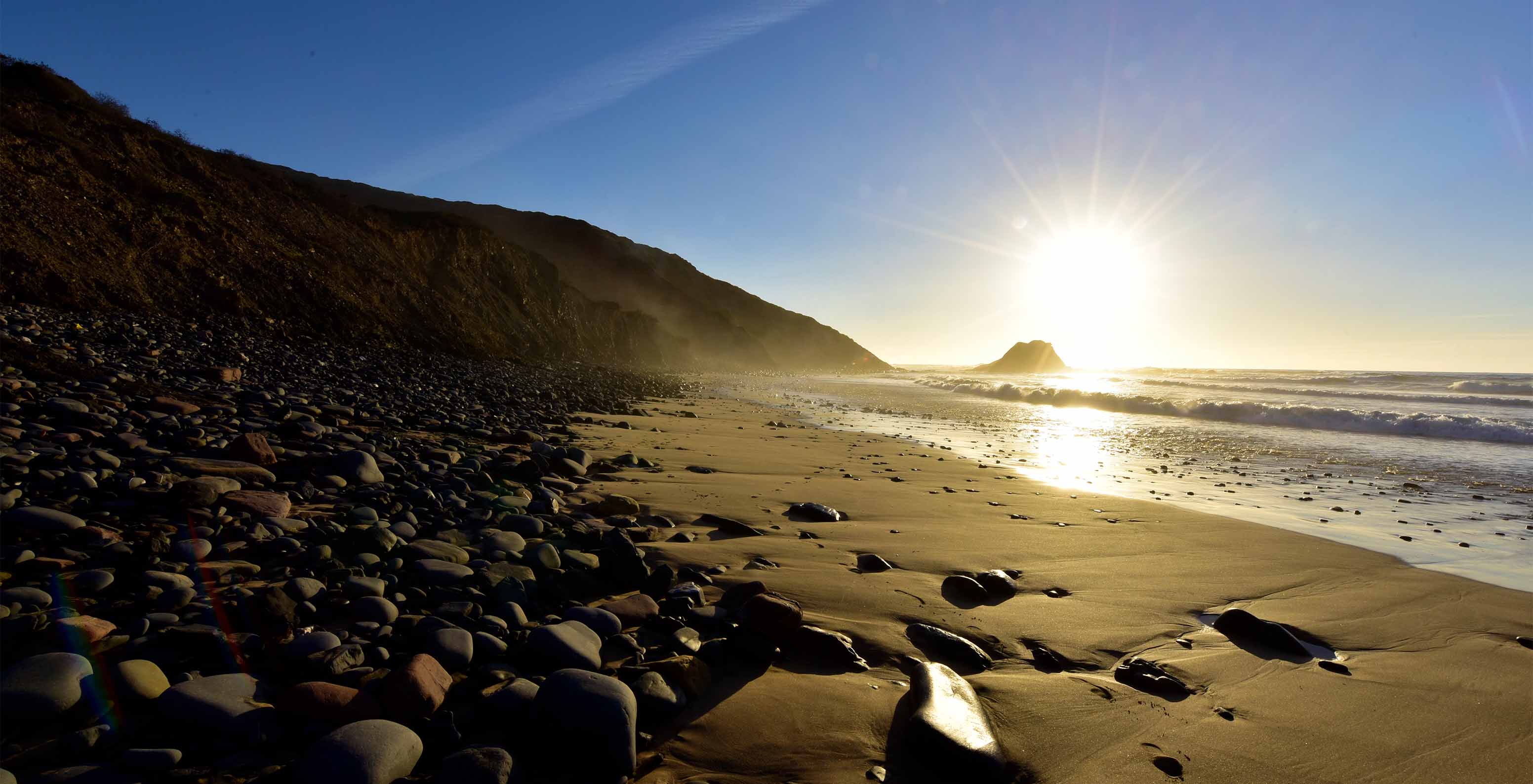 Playa al amanecer, con la luz reflejándose en el mar, piedras en la arena y un acantilado detrás