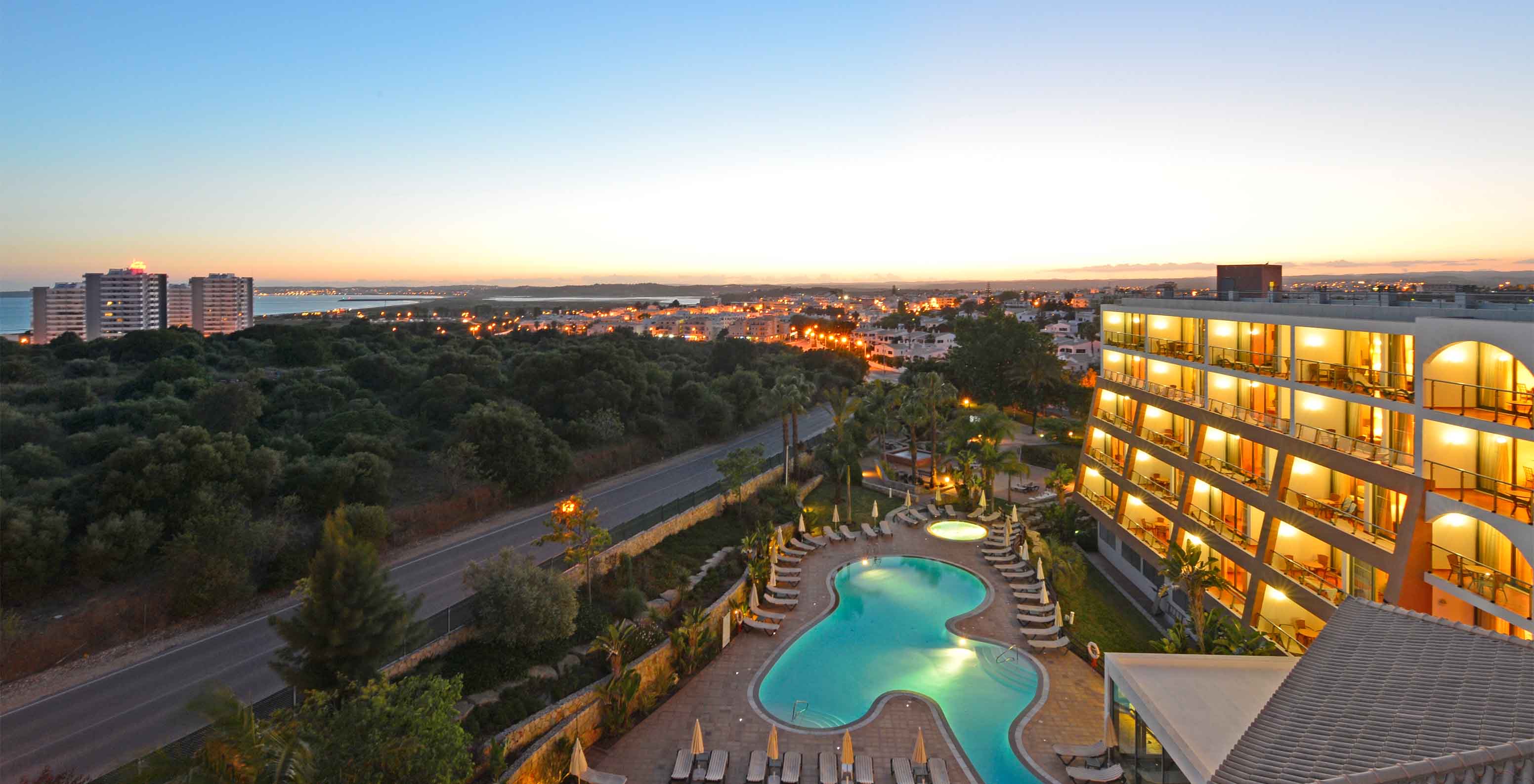 Vista superior del Pestana Alvor Park, con el edificio y la piscina al atardecer, con Alvor al fondo