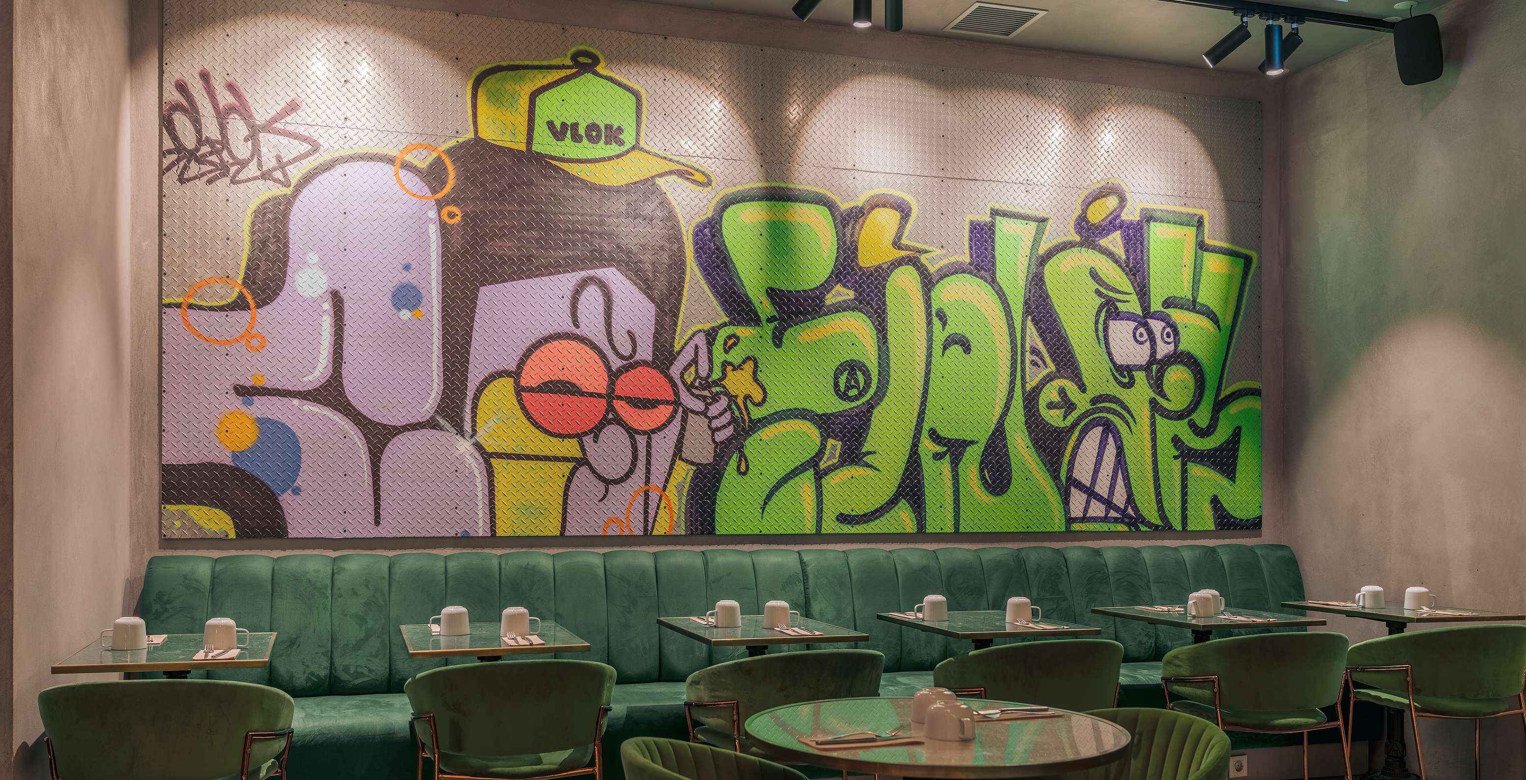 Restaurante con mesas, sillas y sofás verdes, con copas de cerámica blanca y una pared colorida con grafitis