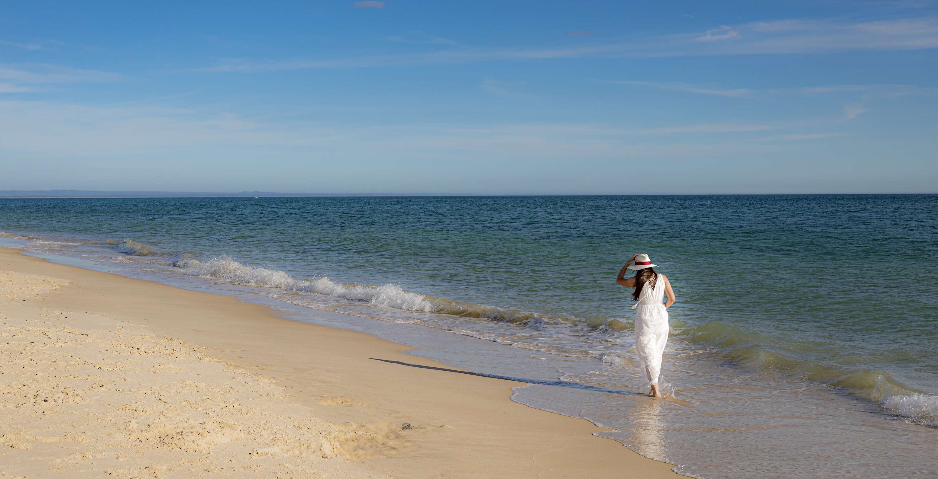 Femme en robe et chapeau blanc se promenant au bord de la mer près du Pestana Tróia Eco-Resort, avec accès direct à la plage
