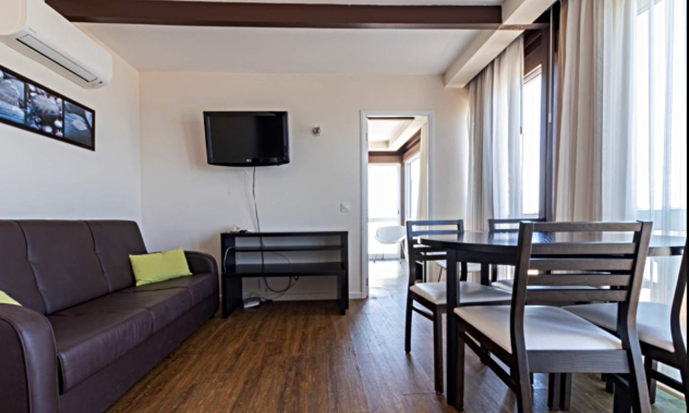 Le Studio Supérieur Vue Mer du Pestana Alvor Atlântico offre un salon lumineux avec canapé, table à manger et télévision, créant une ambiance chaleureuse grâce aux grandes fenêtres laissant entrer une abondante lumière naturelle.