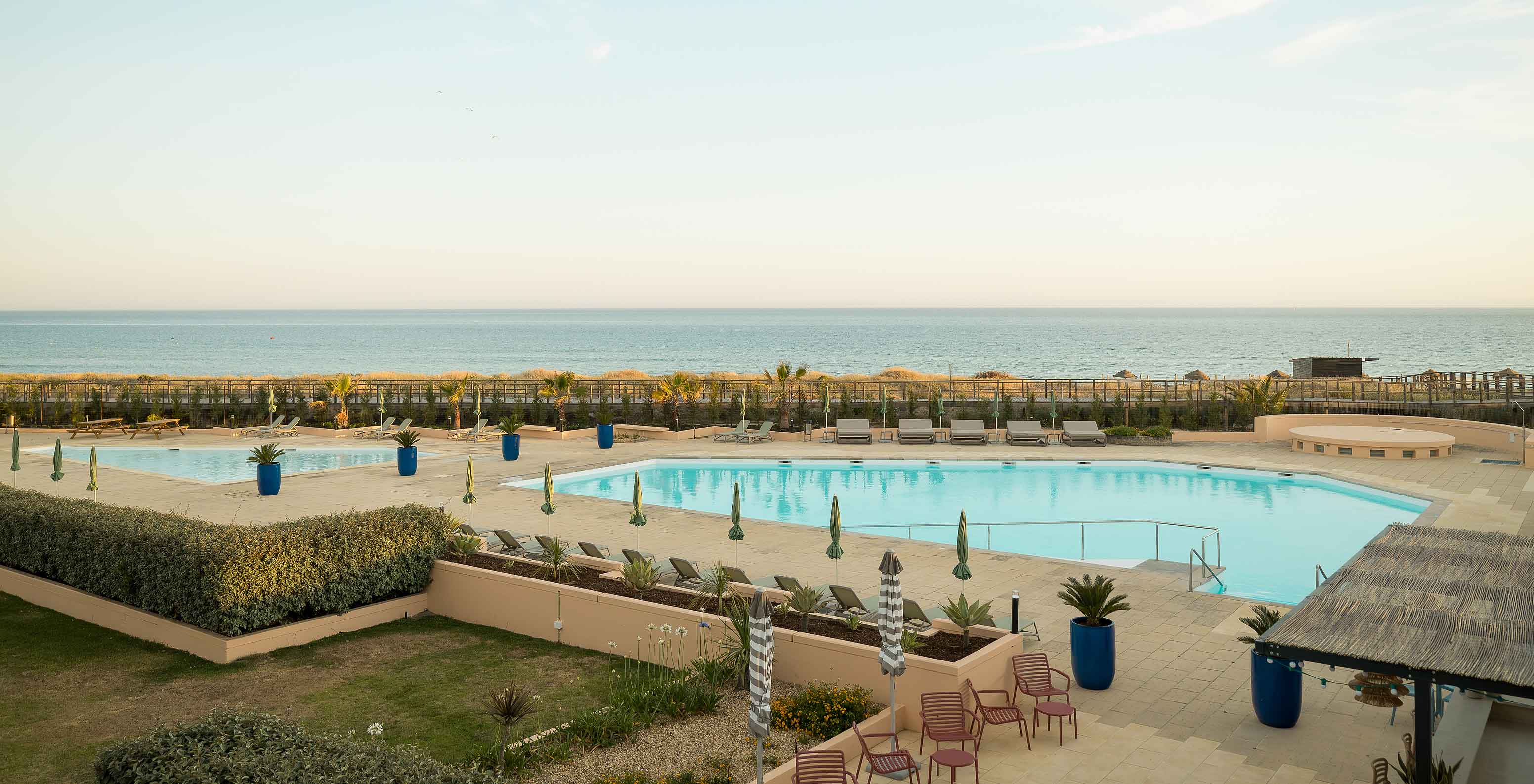 Hôtel près de la plage avec piscine extérieure, plusieurs transats, végétation luxuriante et vue sur la mer bleue