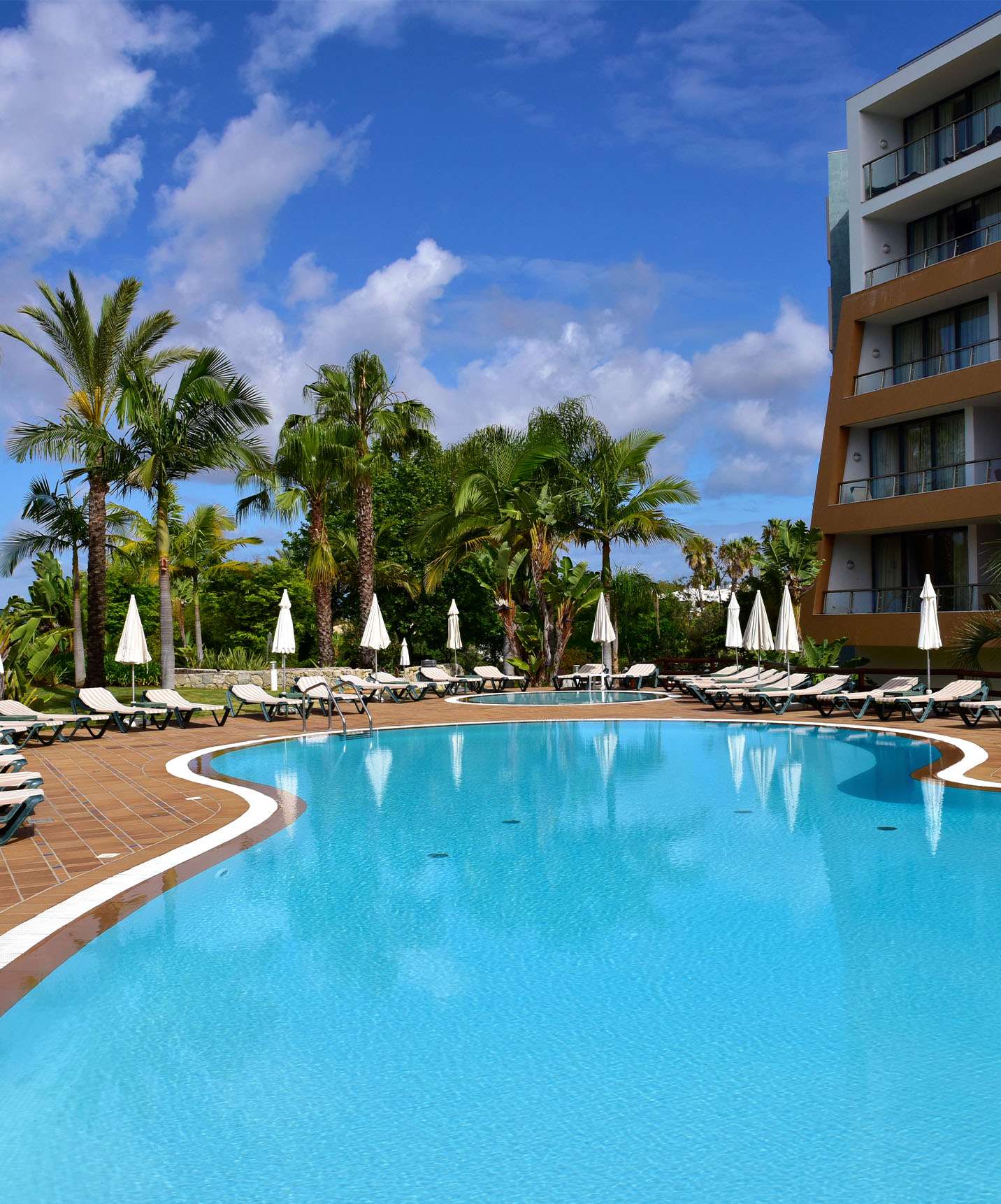 Piscines extérieures avec chaises longues, parasols et palmiers, l'hôtel sur le côté