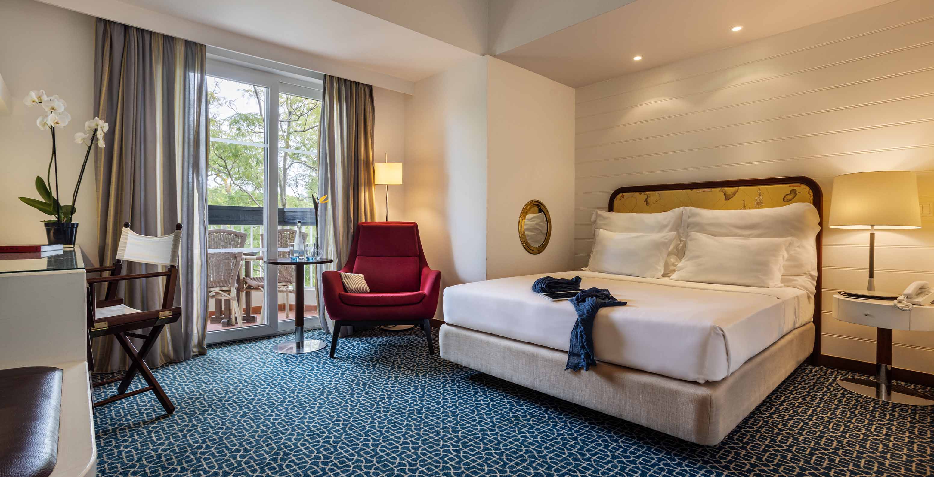 La Suite Vue Jardin du Pestana Alvor Praia a lit double, fauteuil rouge et balcon avec vue jardin et chaises