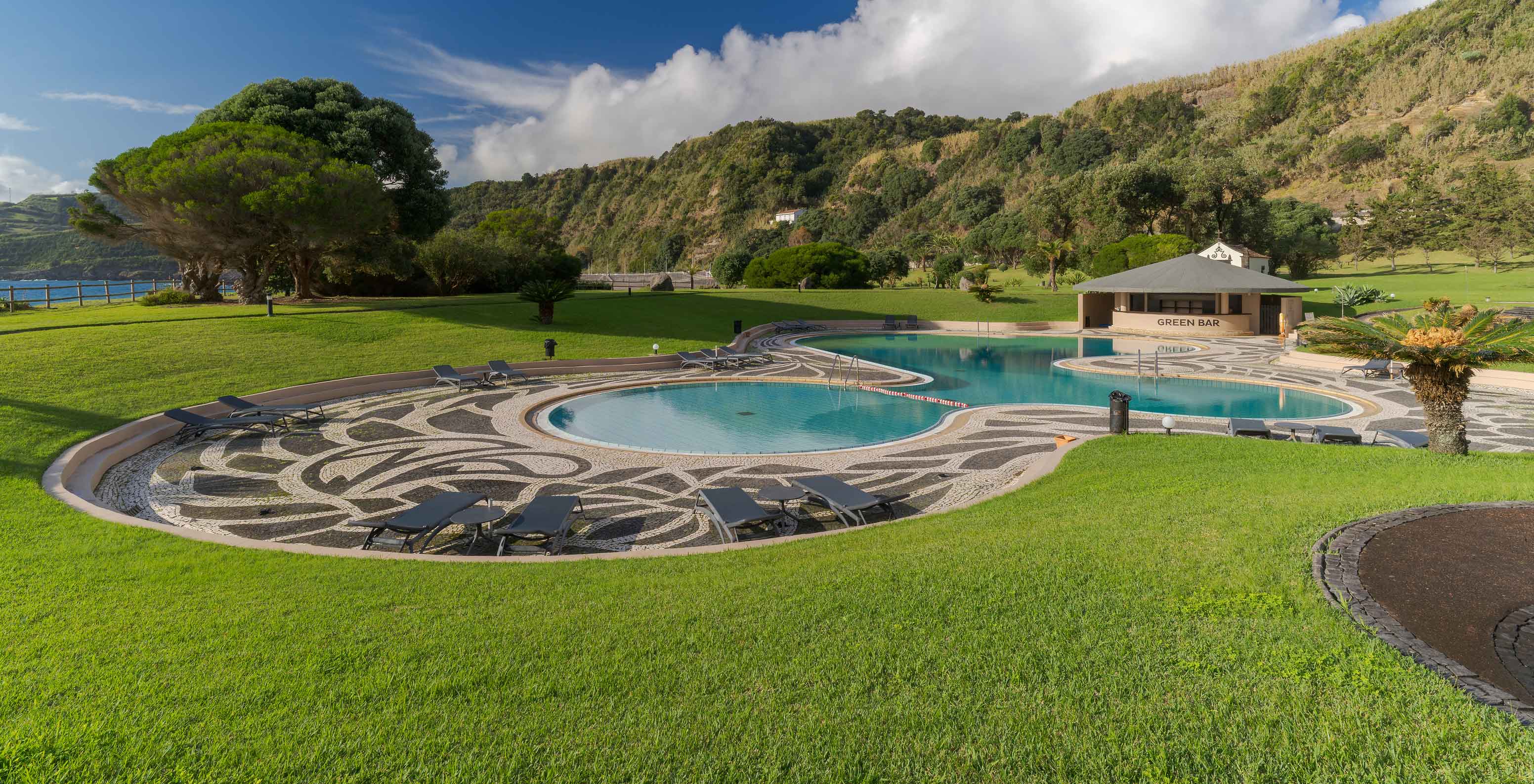 Piscine du Pestana Bahia Praia entourée de jardin et de collines, avec bar au fond et ciel partiellement nuageux