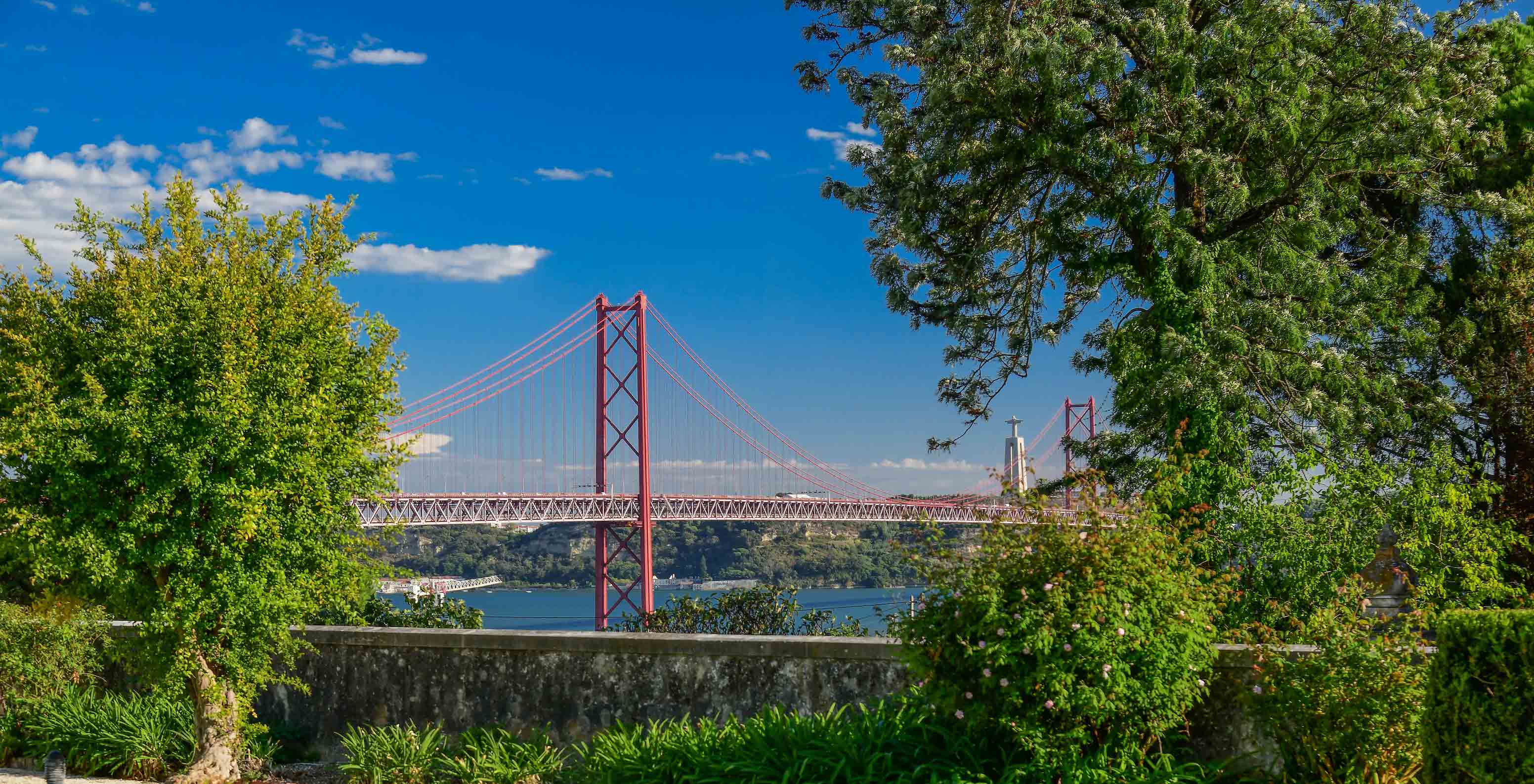 Vue du Pestana Palace Lisbonne vers le pont 25 Avril et le fleuve Tage, avec plusieurs bâtiments et arbres entre