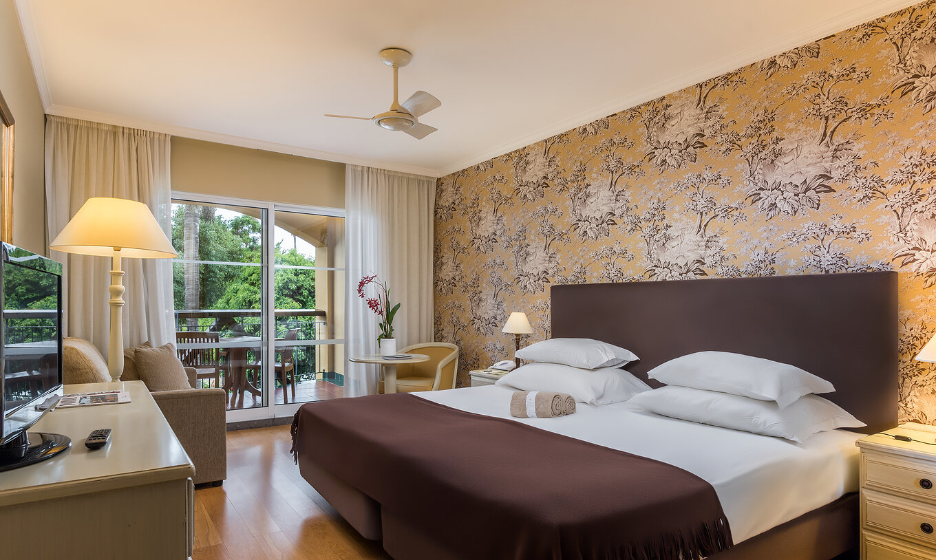 Le Studio Classic du Pestana Village a un lit double avec tête de lit et couvre-lit marron, papier peint floral et balcon