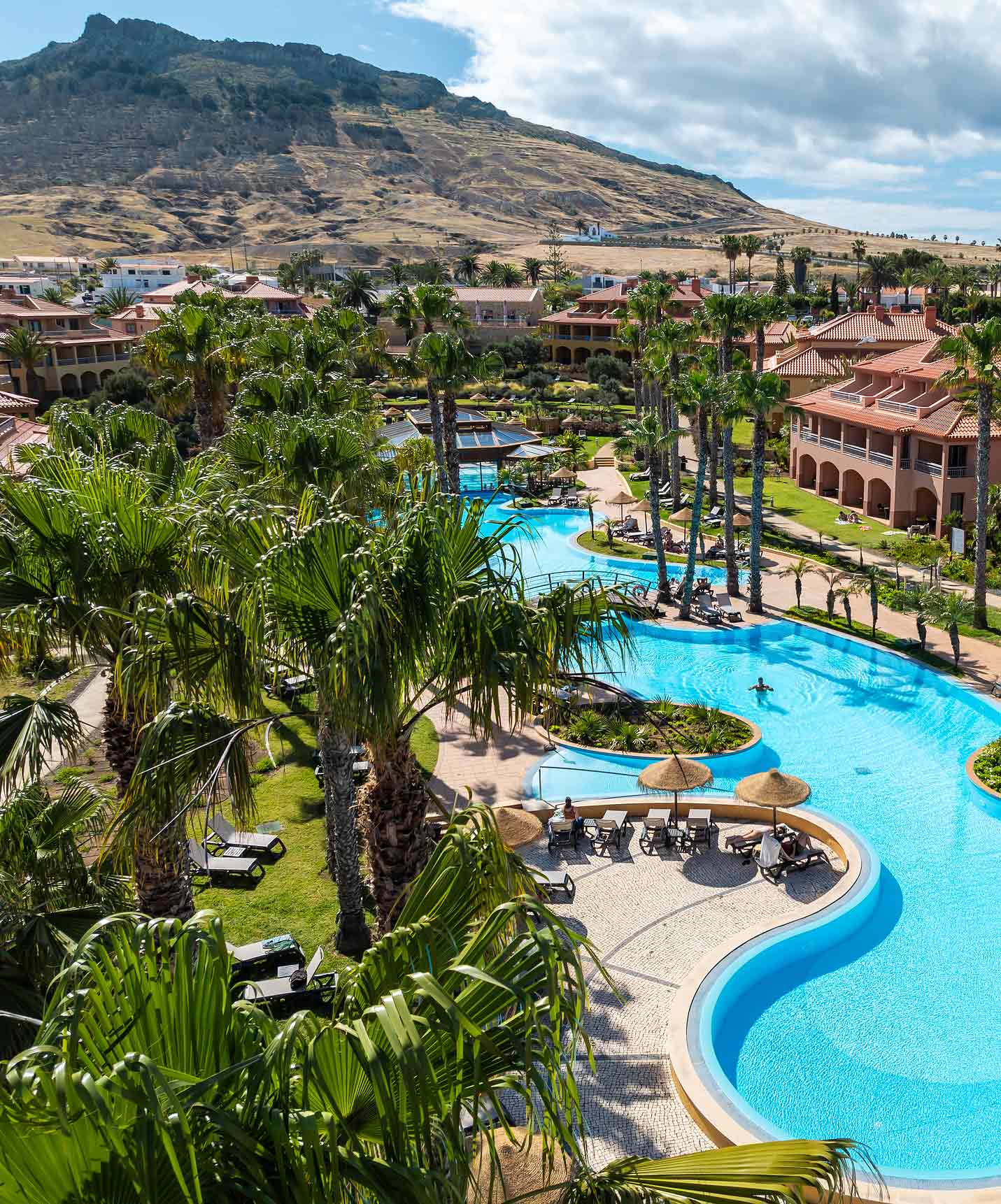 Vue de la piscine extérieure du Pestana Porto Santo All Inclusive, entourée de jardin, de palmiers et de chaises longues.