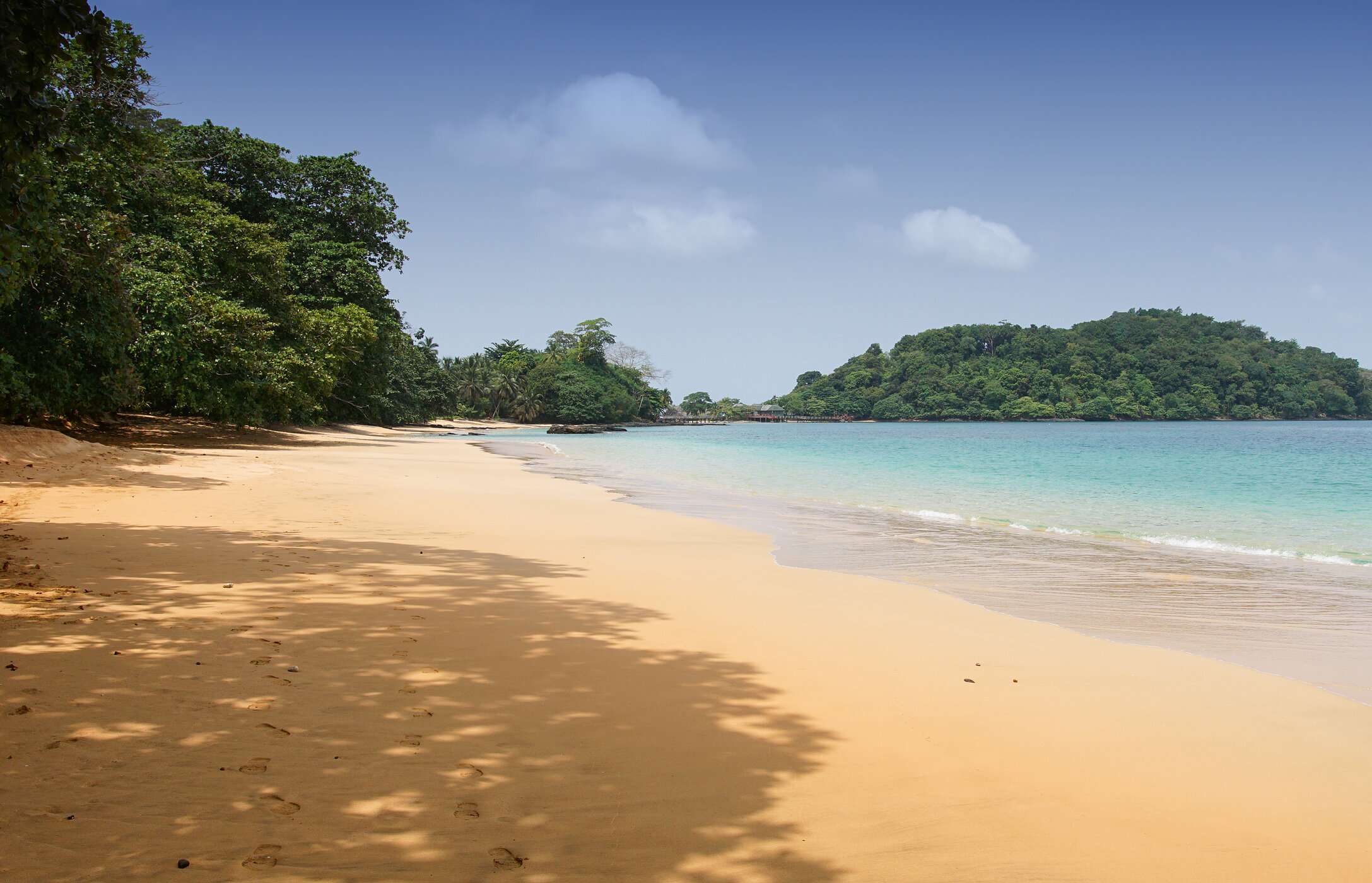 Vue de la plage de Coco à São Tomé et Príncipe, avec du sable doré, des eaux calmes cristallines et de la végétation autour.