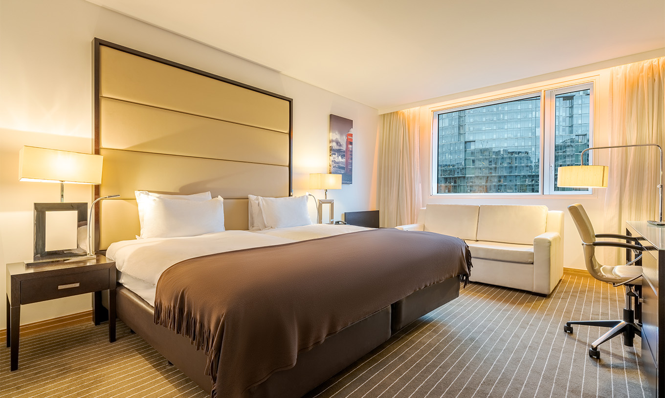 Le Twin Deluxe du Pestana Chelsea Bridge a deux lits, table de nuit, canapé, lampes et fenêtres avec rideaux