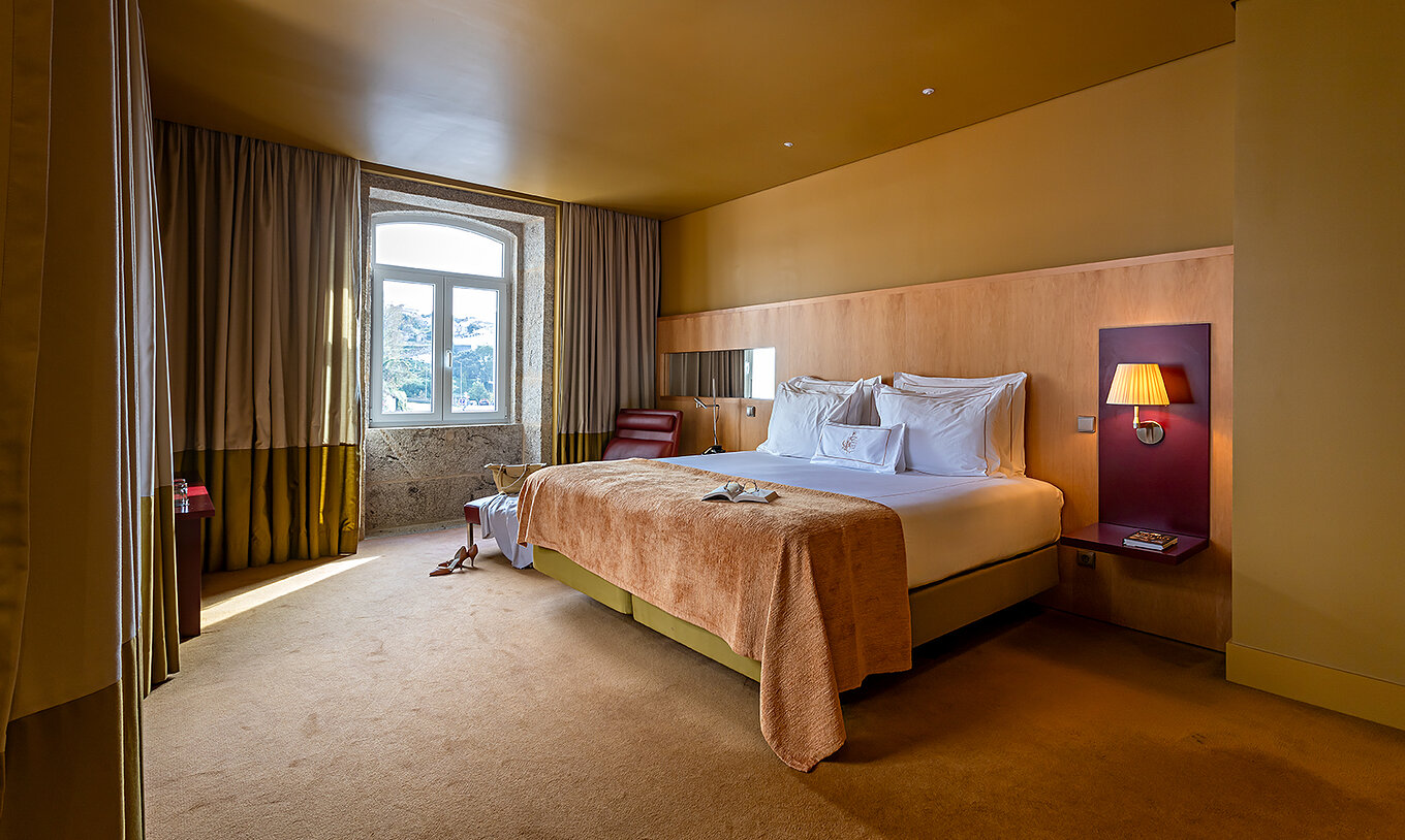 De Junior Suite heeft een tweepersoonsbed met beige deken en een stenen raam met uitzicht op de rivier