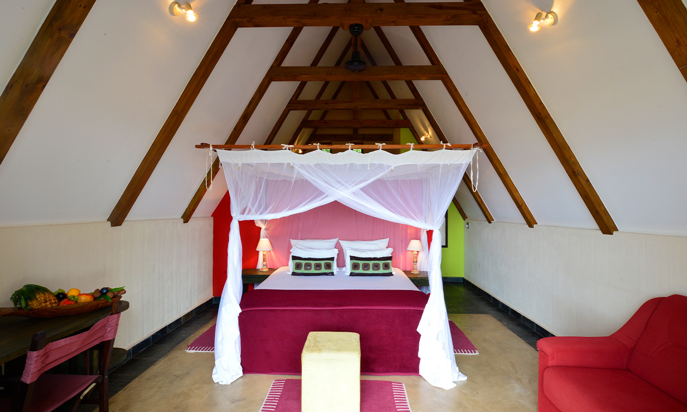 O Bungalow T1 do Pestana Bazaruto Lodge tem uma cama de casal com dossel, um sofá e uma secretária com uma taça com frutas