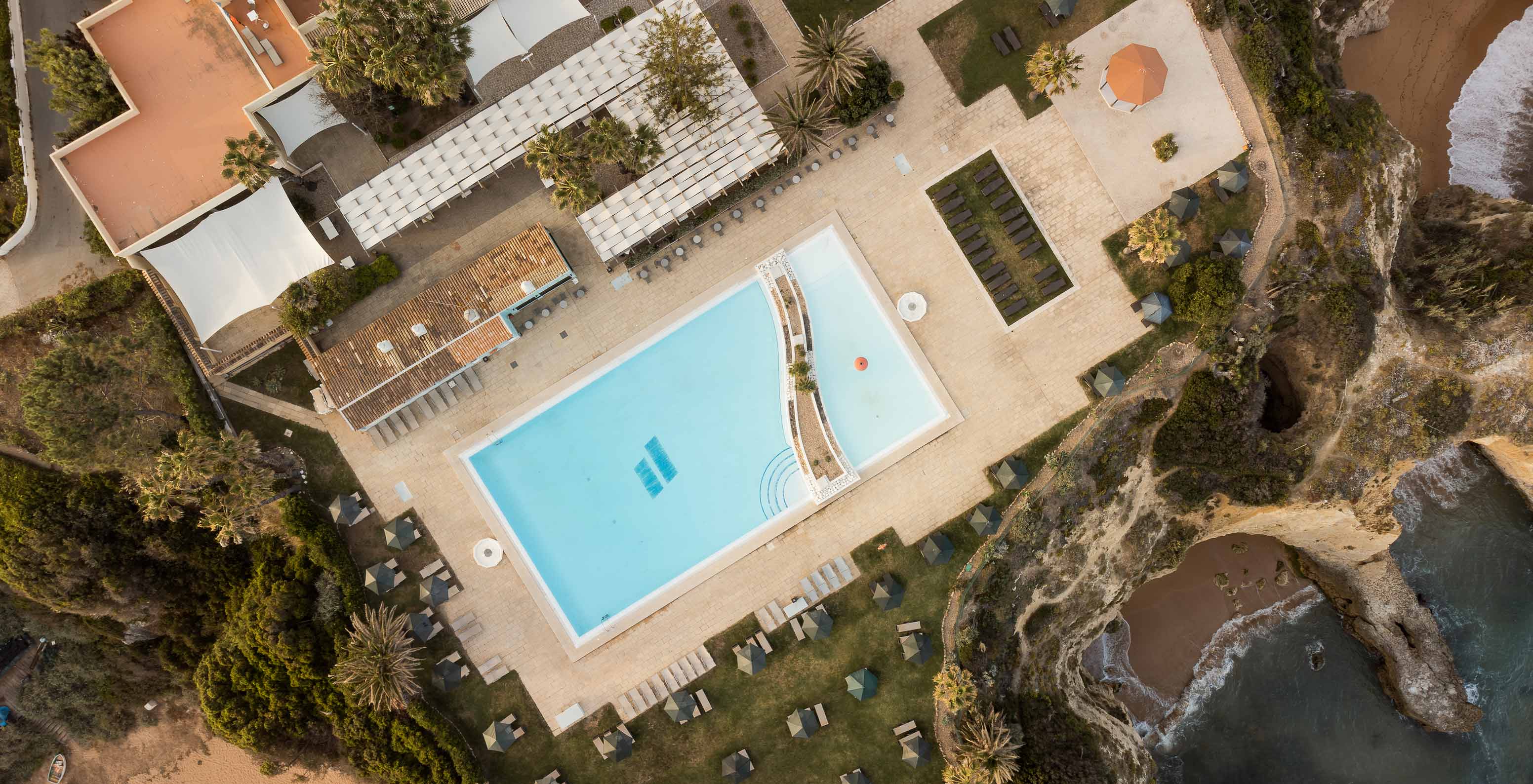 Vista de drone da piscina exterior do Pestana Viking, com várias palmeiras, espreguiçadeiras e com vista para o mar