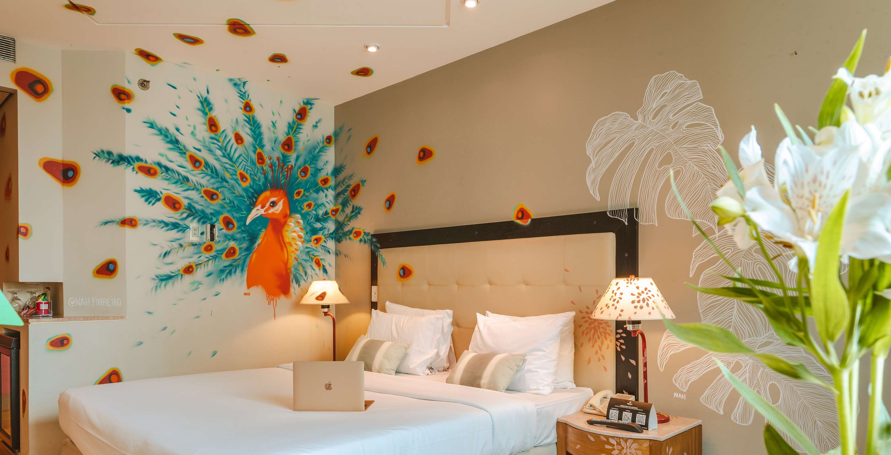 Quarto decorado pela artista Nah Pinheiro, com um pavão colorido na parede, em hotel 4 estrelas em São Paulo