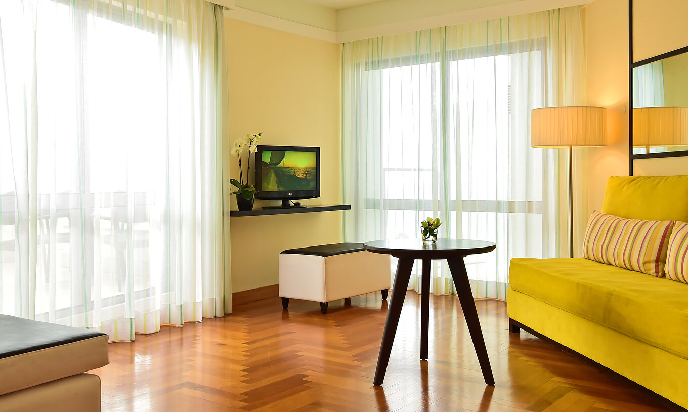 A Junior Suite do Pestana Promenade tem uma sala de estar com um sofá verde, uma televisão e duas janelas grandes