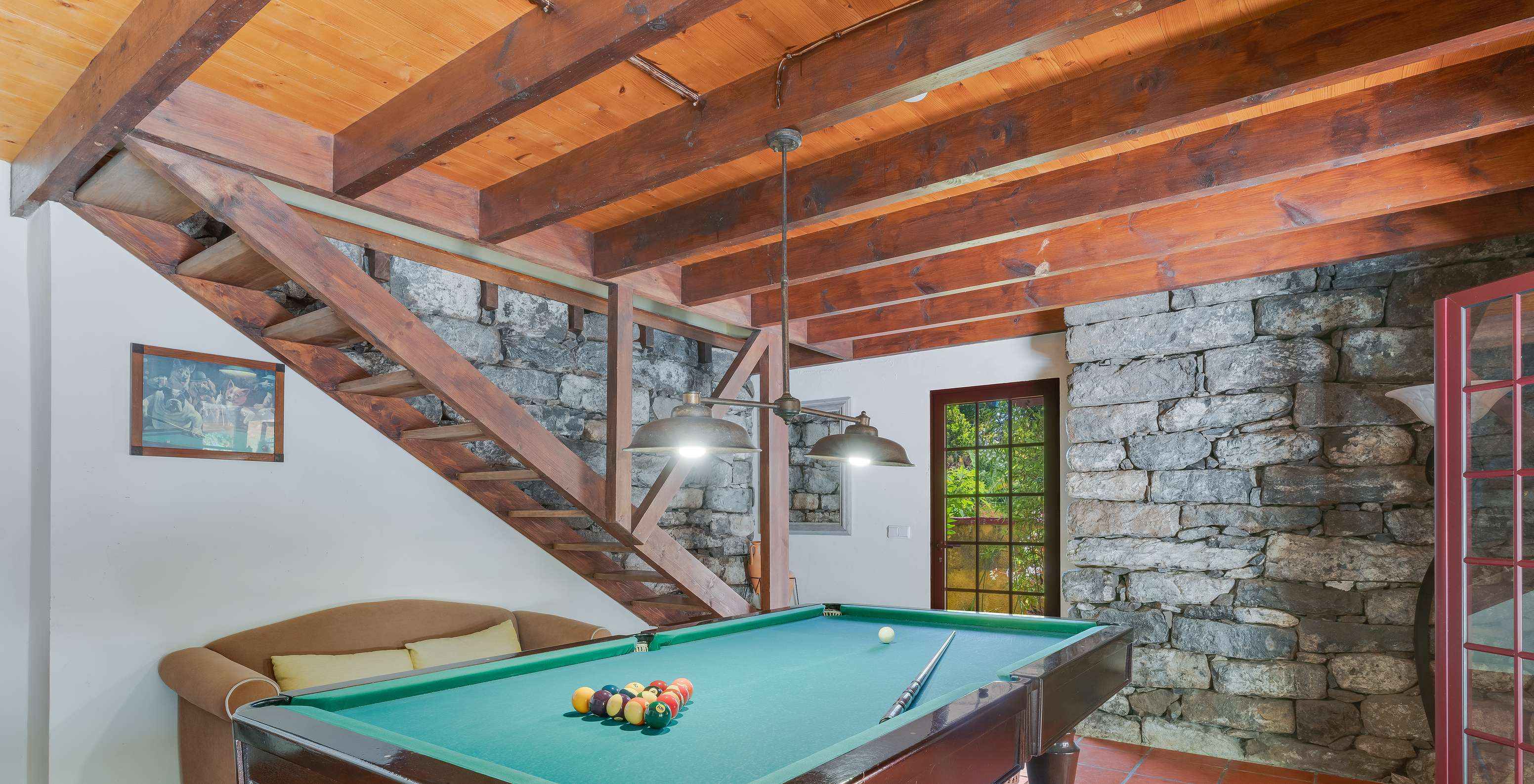 Sala de jogos com mesa de snooker, com sofá, escadas de madeira e paredes de pedra, com portas e janelas para o exterior