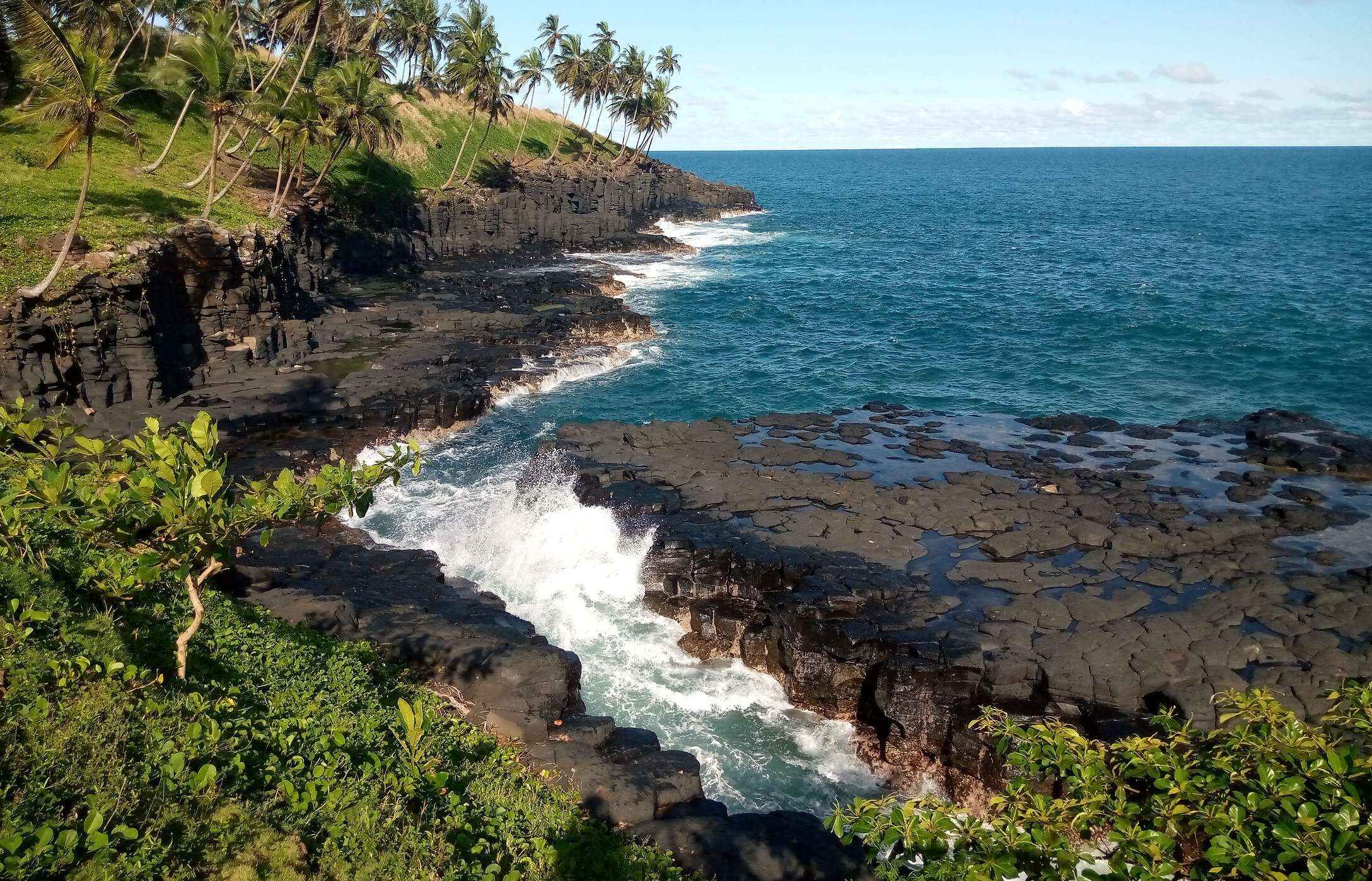 Vista para a Boca do Inferno em São Tomé e Príncipe, com mar agitado, rochas negras e vegetação em redor