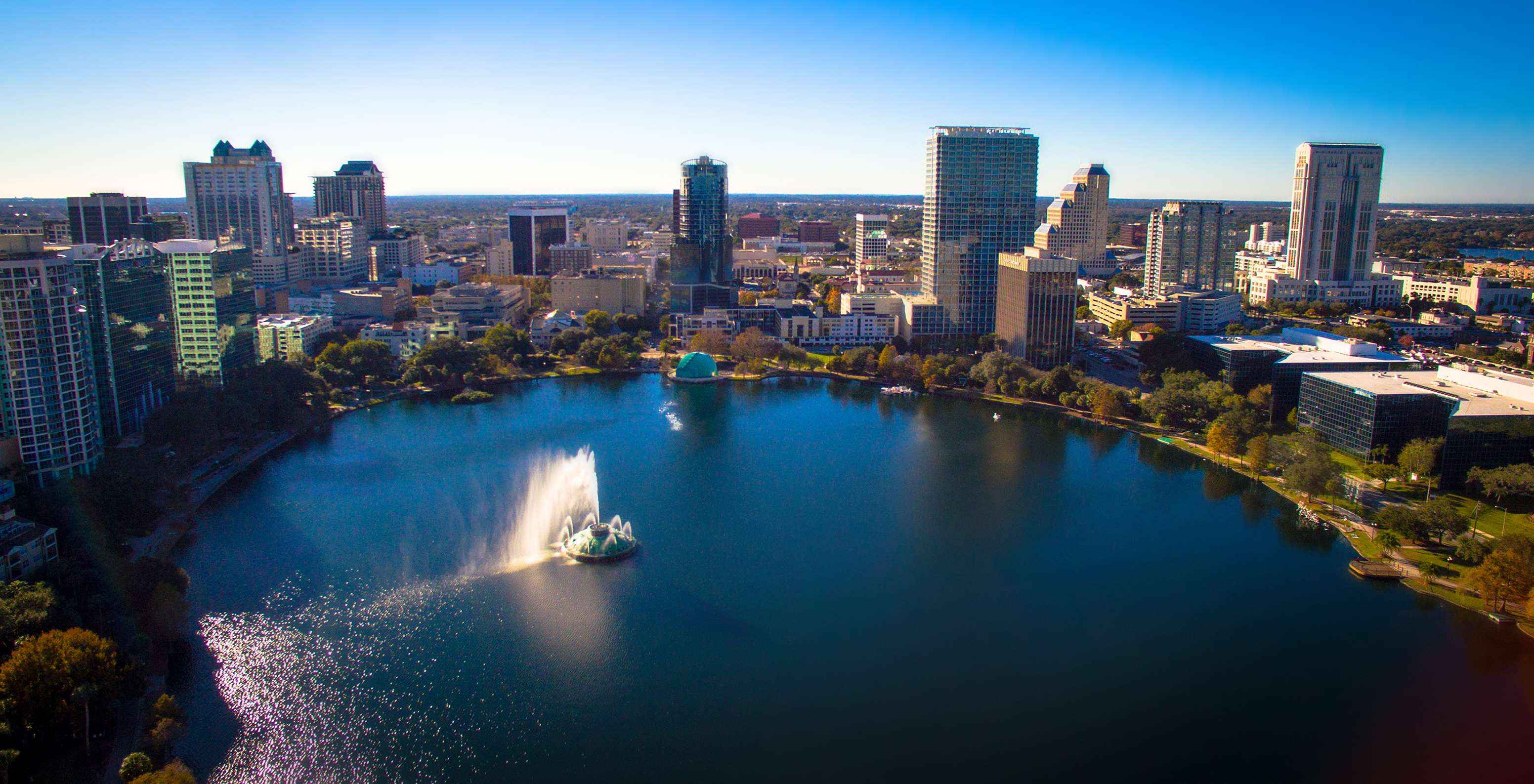Vista aérea do centro de Orlando, num dia de céu azul com poucas nuvens, com o Lago Eola e seus edifícios icônicos ao fundo