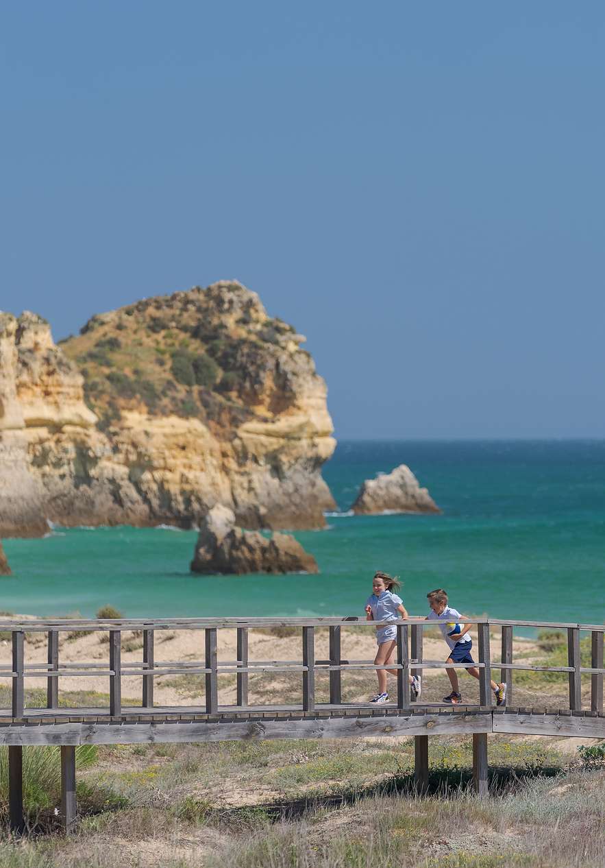 Eltern und zwei Kinder gehen auf einem Holzsteg mit Blick auf das hellblaue Meer und die Klippen im Hintergrund