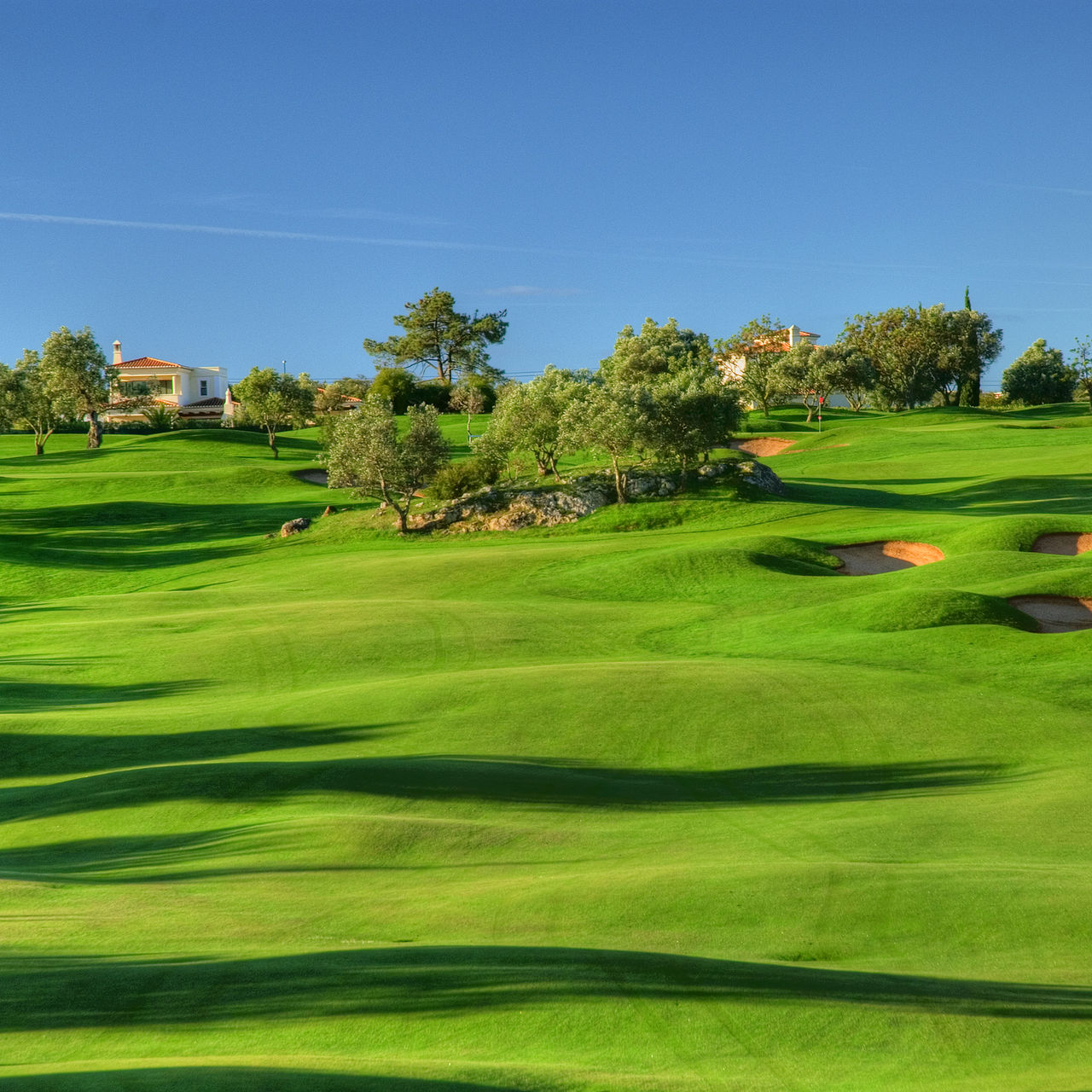 Üppiger Golfplatz in hügeligem Gelände bei Gramacho, blauer Himmel, Bäume, Büsche und einige Häuser im Hintergrund