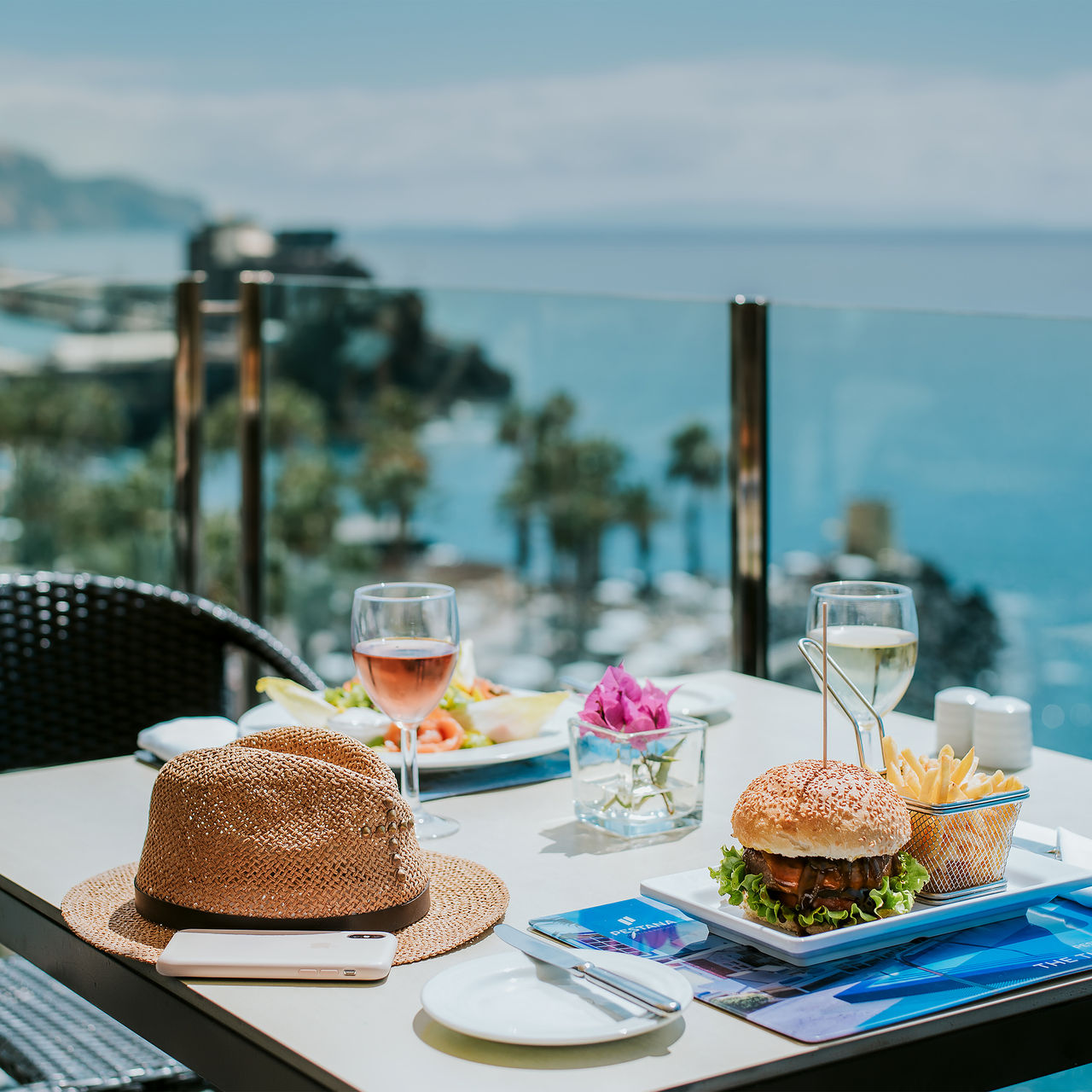 Tisch im Pestana Madeira Beach Club mit Meerblick, darauf Hamburger, Pommes, Wein und ein Hut