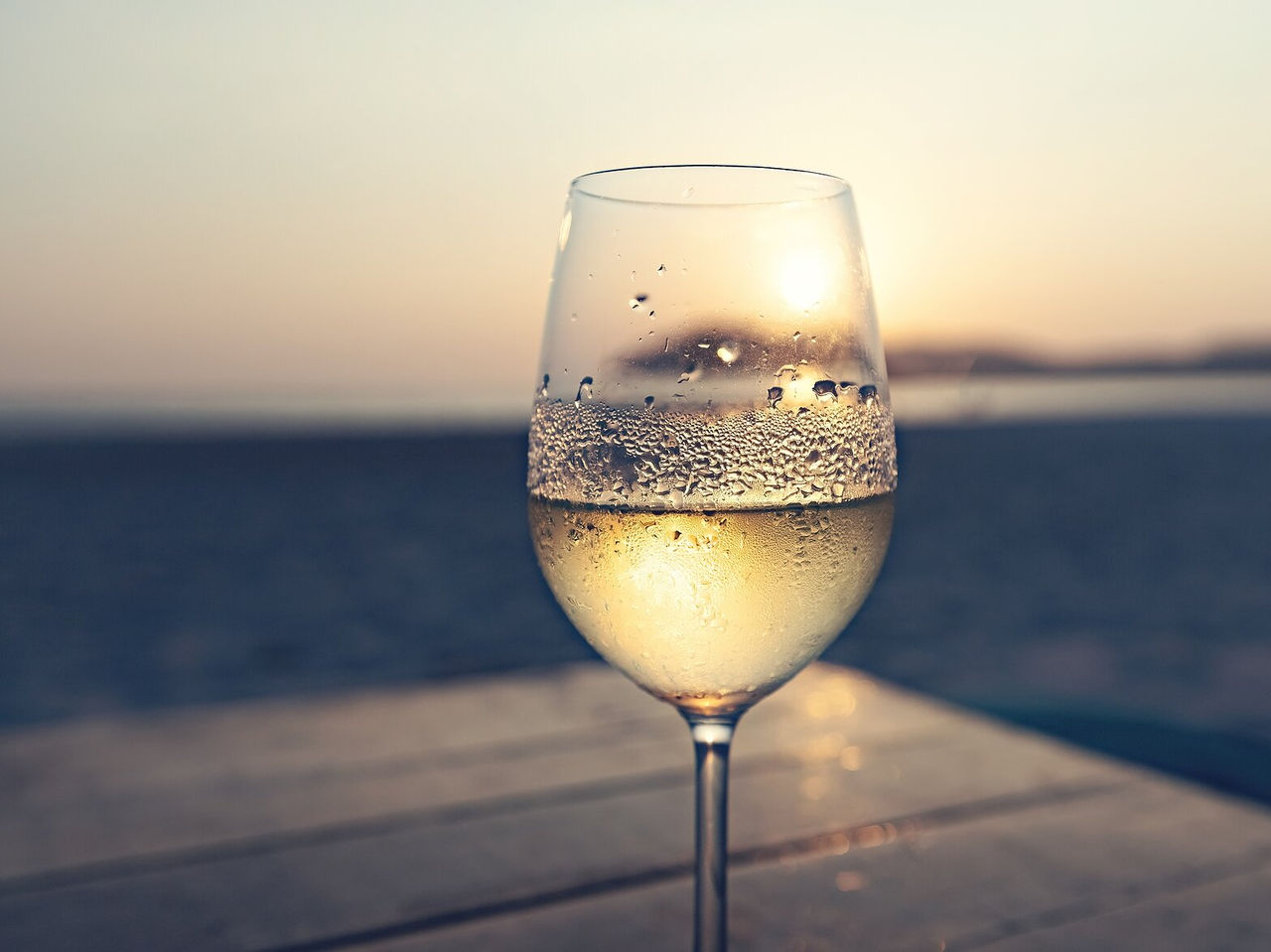 Glas Weißwein mit unscharfen Sonnenuntergangsreflexionen im Hintergrund, auf einer Holzoberfläche