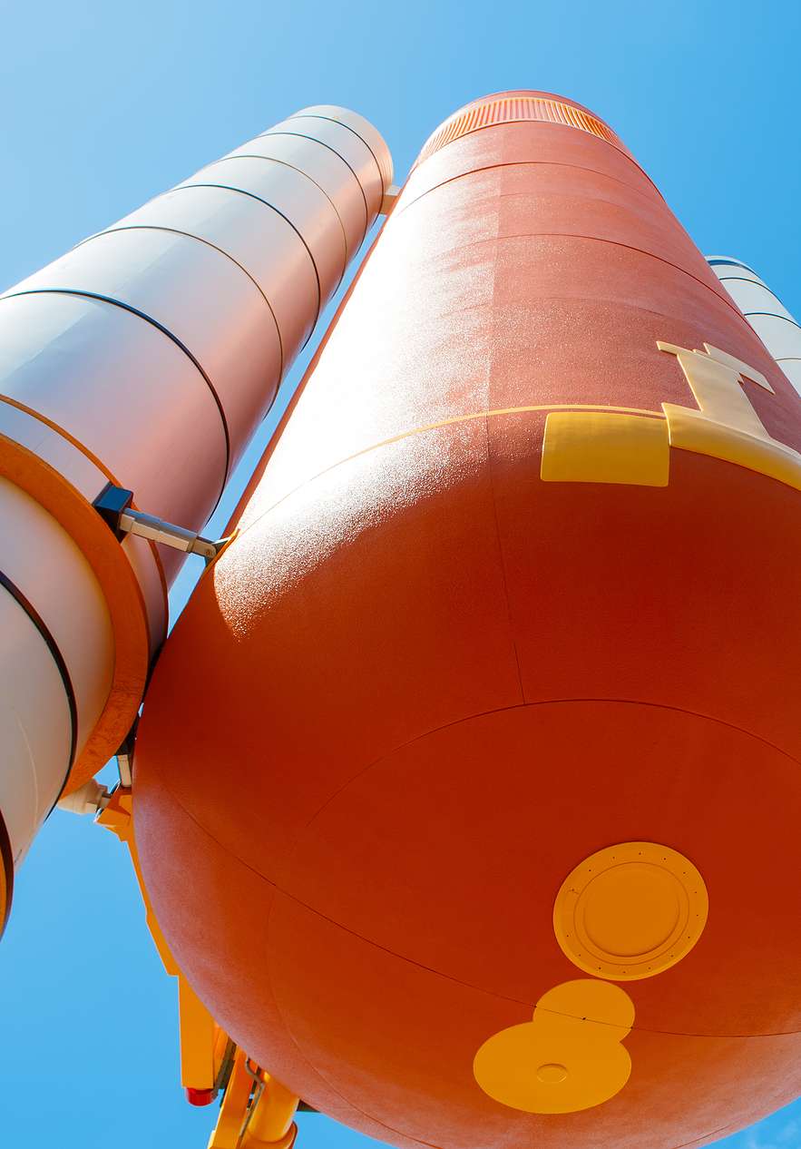 Blick von unten auf einen externen Tank einer orange-weißen Rakete, mit blauem Himmel und einigen Wolken im Hintergrund