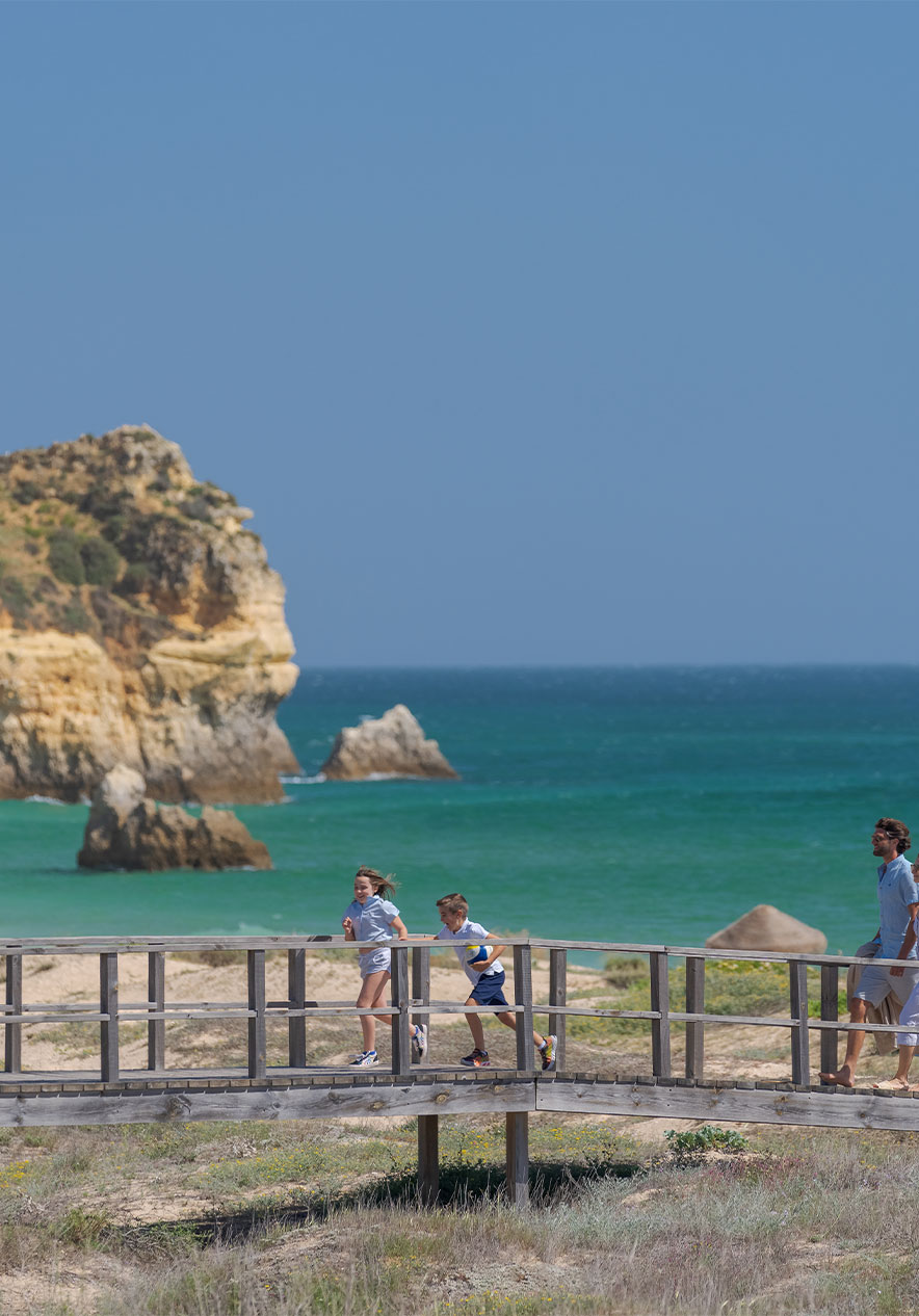 Children run smiling on the boardwalk of Alvor beach where we see crystal blue water and golden sand