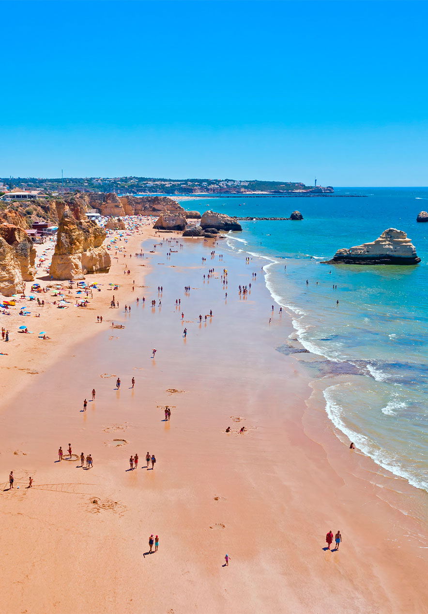 Vista aérea de la Playa da Rocha, con sombrillas, acantilados, personas y mar, bajo un cielo despejado