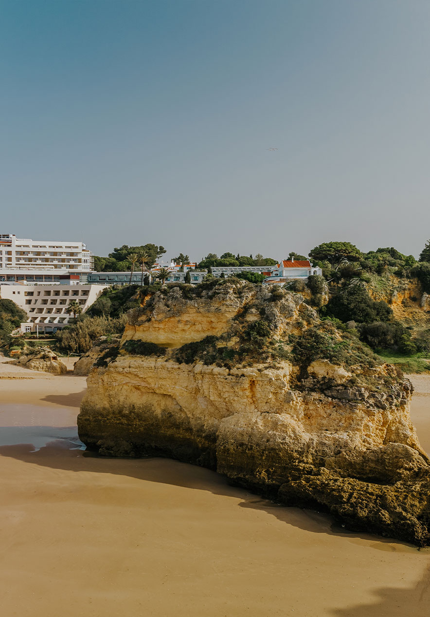 Playa de Alvor con rocas, arena, vegetación, acantilado y edificios al fondo, bajo cielo despejado
